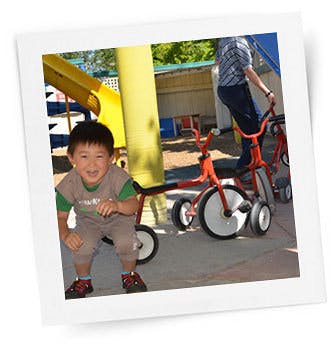 Child crouching next to a tricycle, person on another tricycle in background