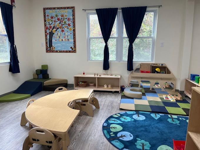 A classroom with small tables and chairs, play mats, and a colorful bulletin board