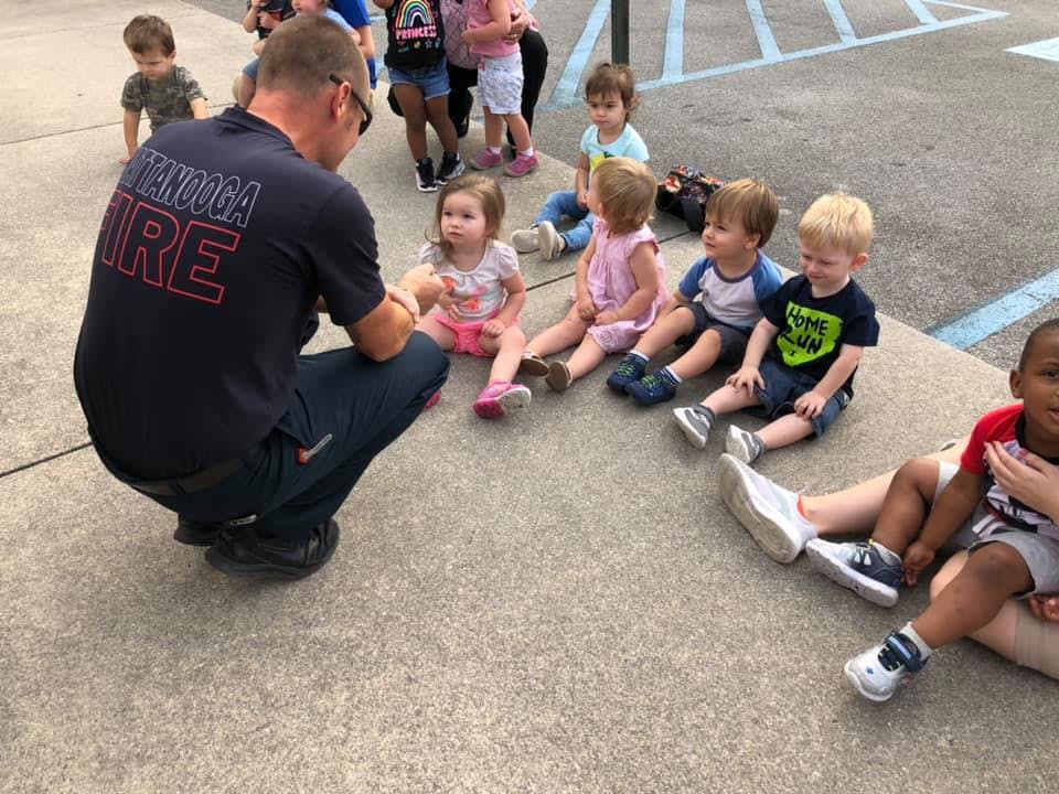 Adult in a fire department shirt crouching with children sitting on the ground