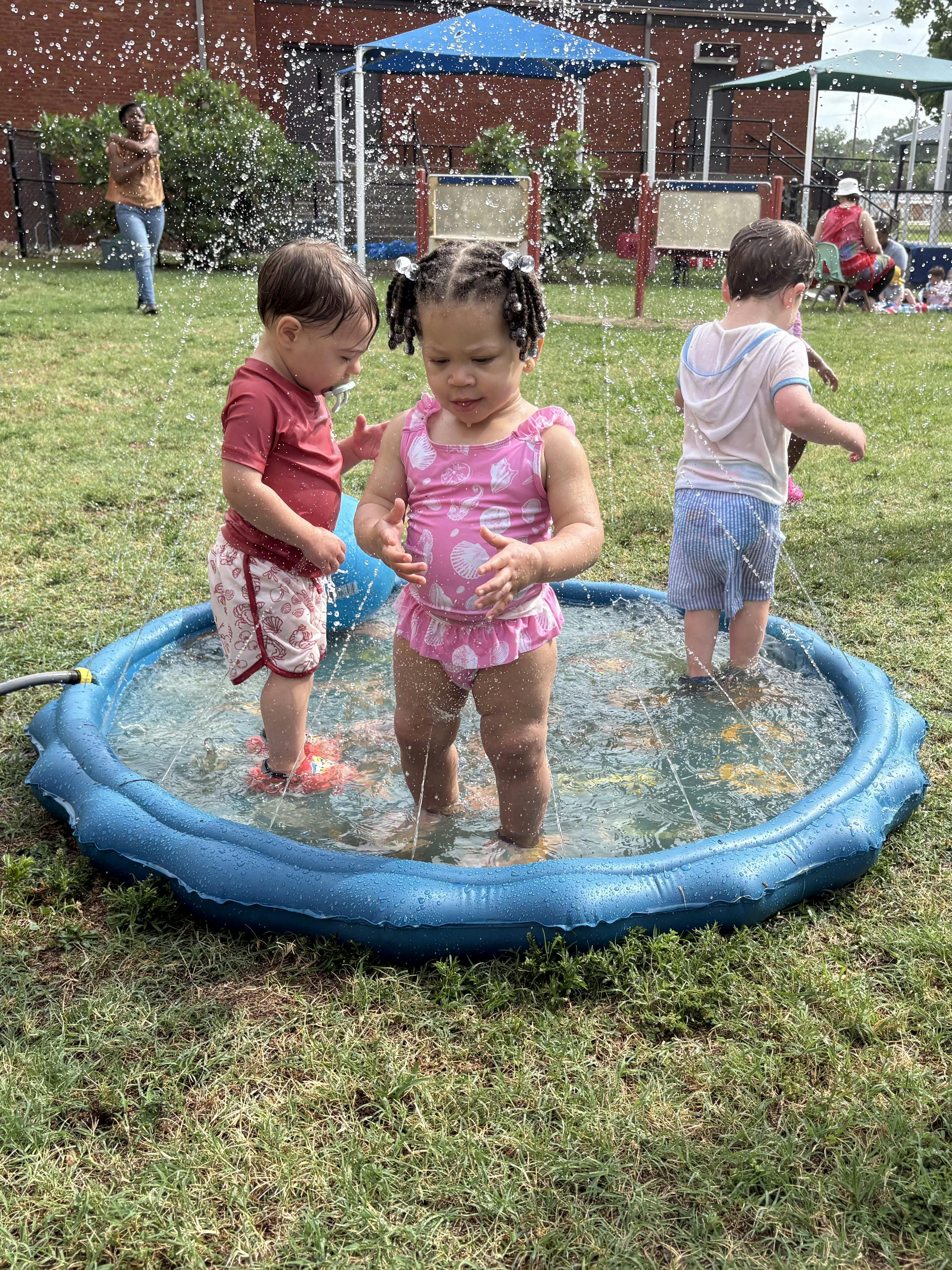 Three children playing in a shallow inflatable pool with water sprayers
