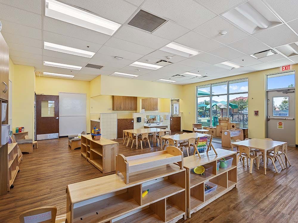 Empty classroom with small wooden tables and chairs, shelves, and a window overlooking a playground
