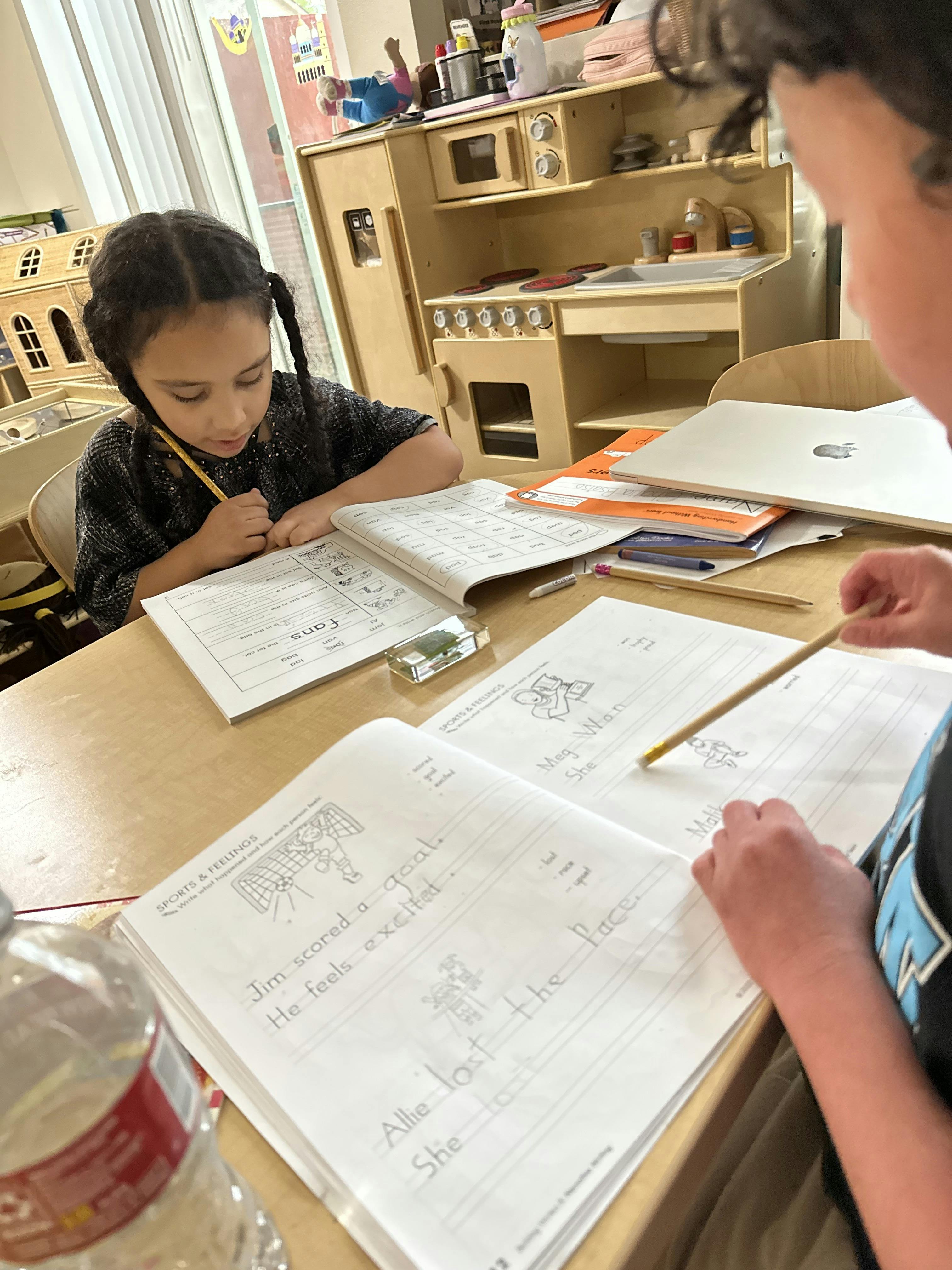 Two children writing in notebooks at a table in a classroom