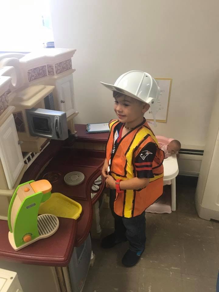 Child in a construction vest and hard hat standing next to a play kitchen