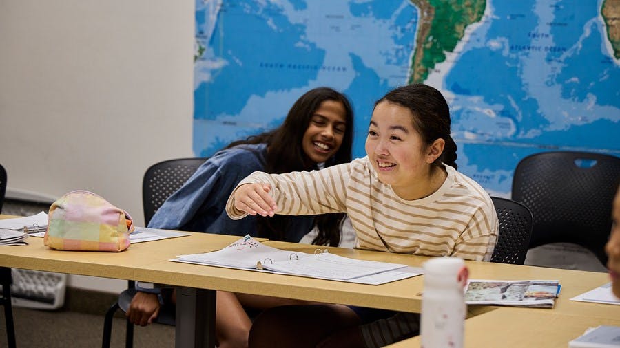 Two teens sitting at a classroom table with a world map behind them
