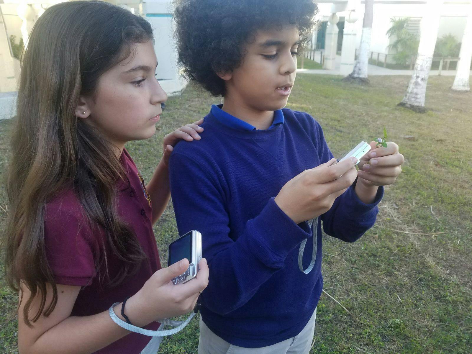Two children holding small devices and looking at a plant