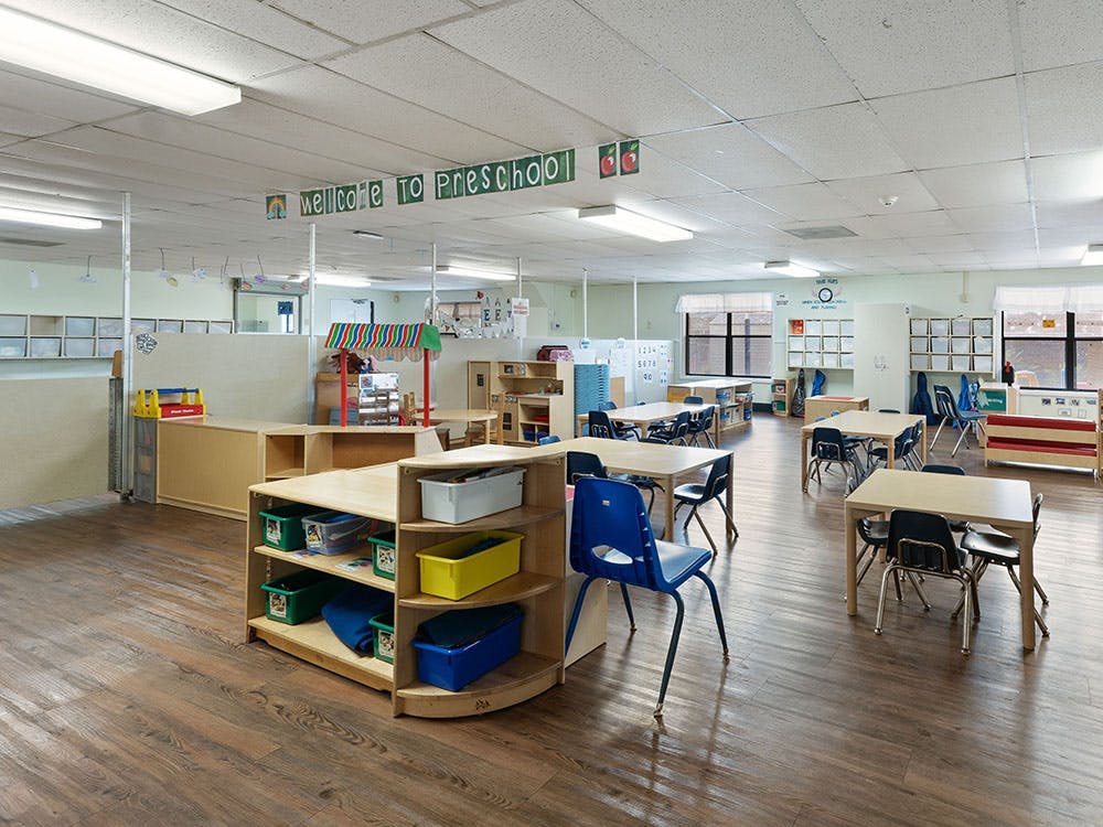 A classroom with tables and chairs, shelves, and a banner reading "WELCOME TO PRESCHOOL"