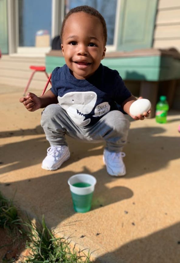 Child crouching outdoors holding an egg, with a cup of green liquid on the ground