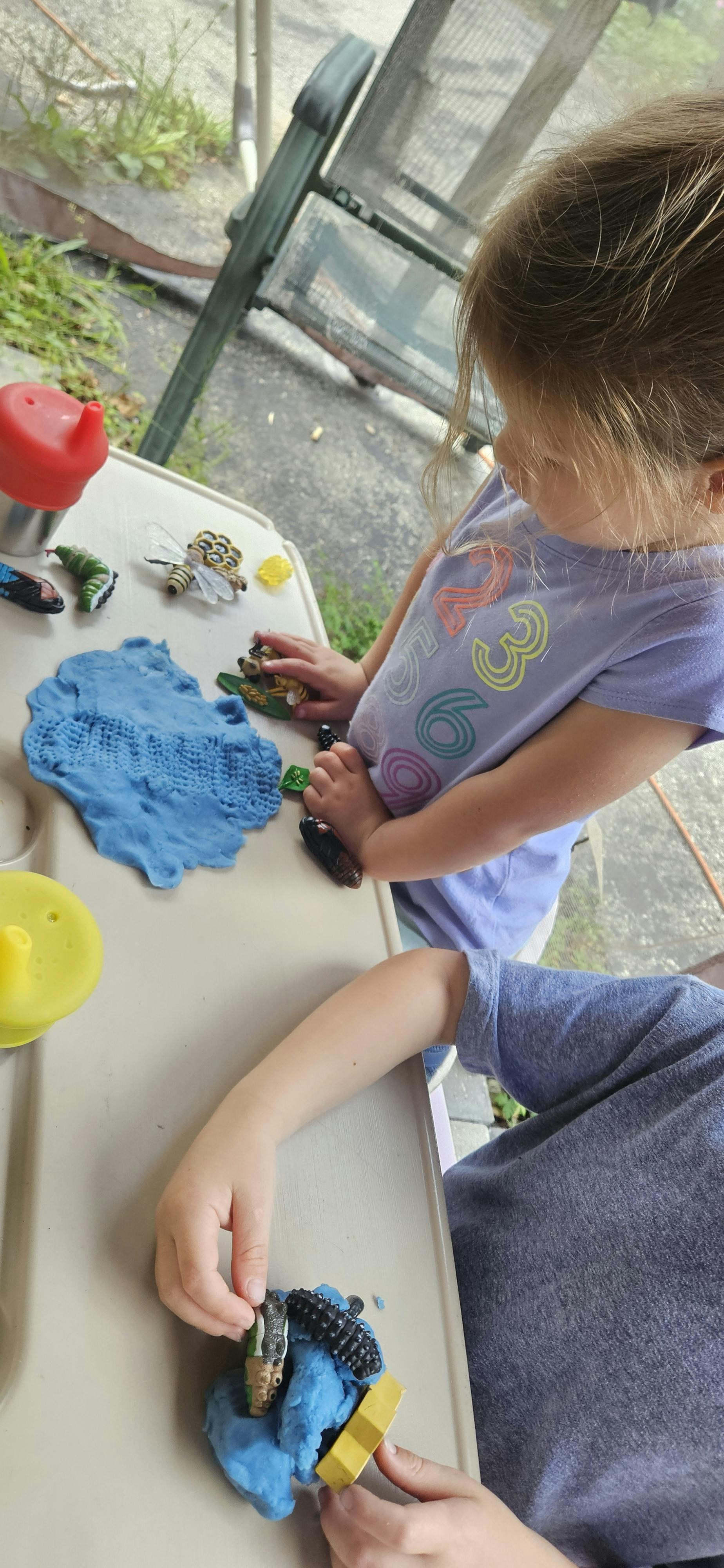 Two children playing with blue playdough and small toys on a table