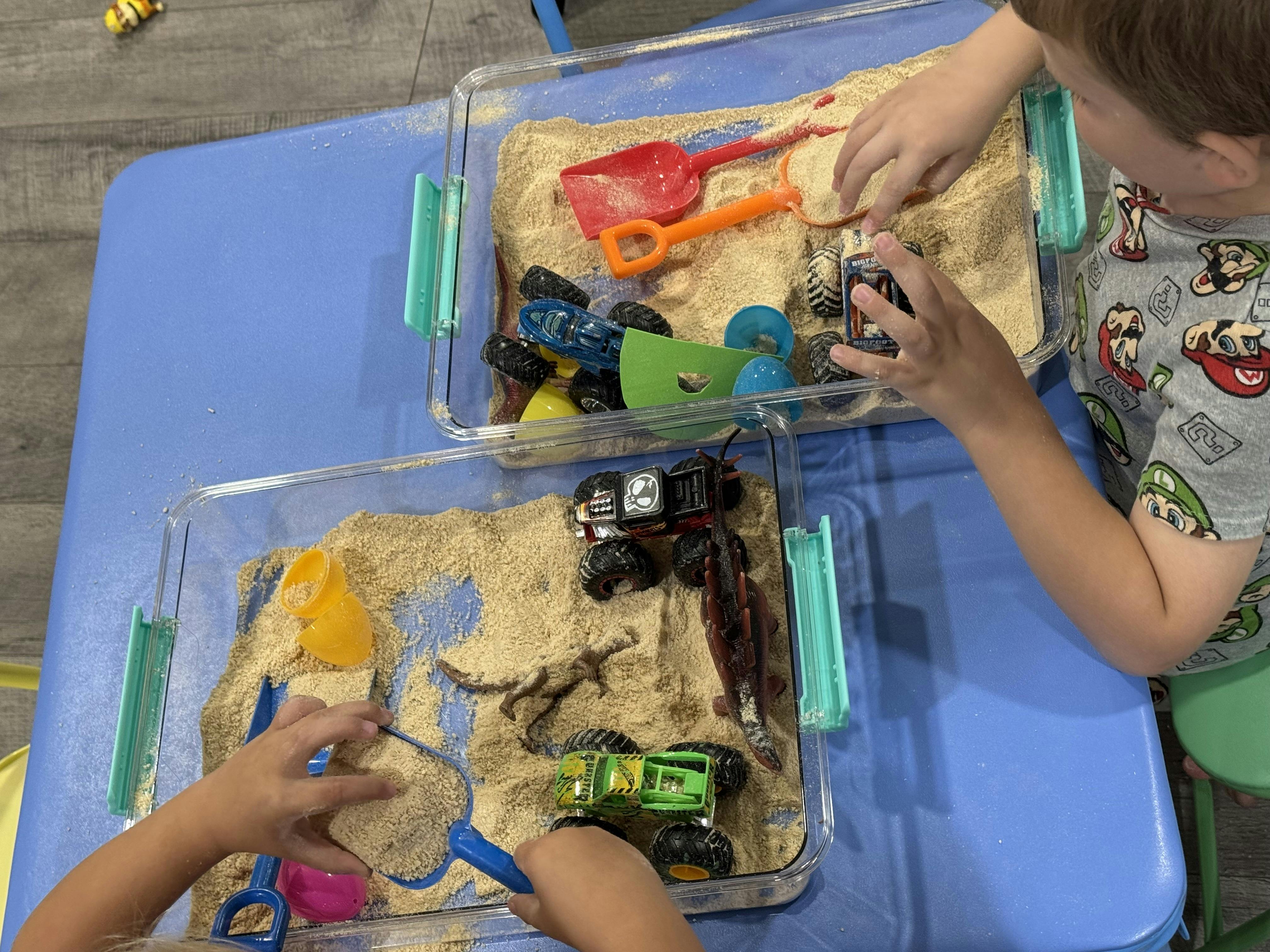 Two children playing with sand and toy trucks in clear bins