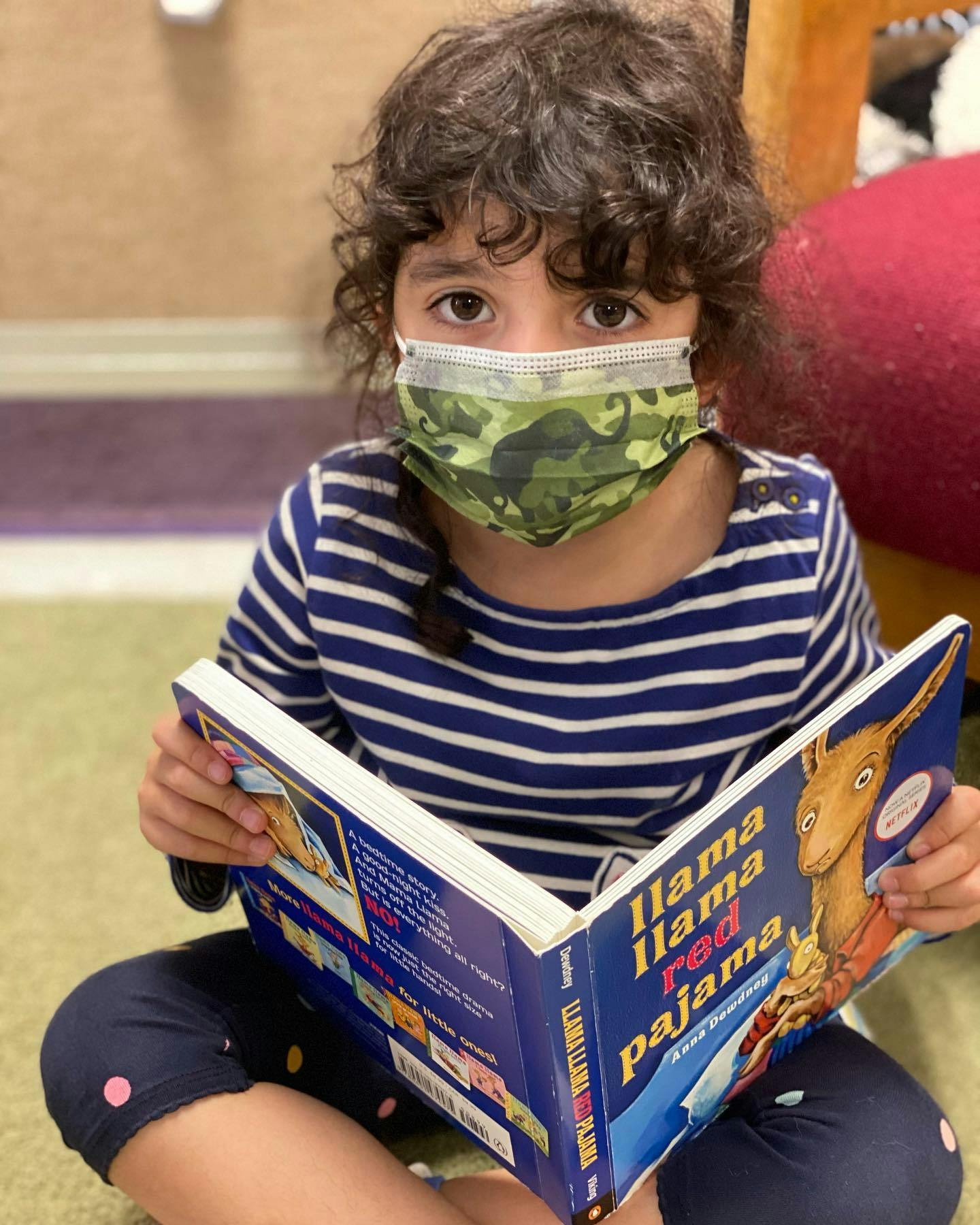 Child in a face mask reading a book titled "Llama Llama Red Pajama"