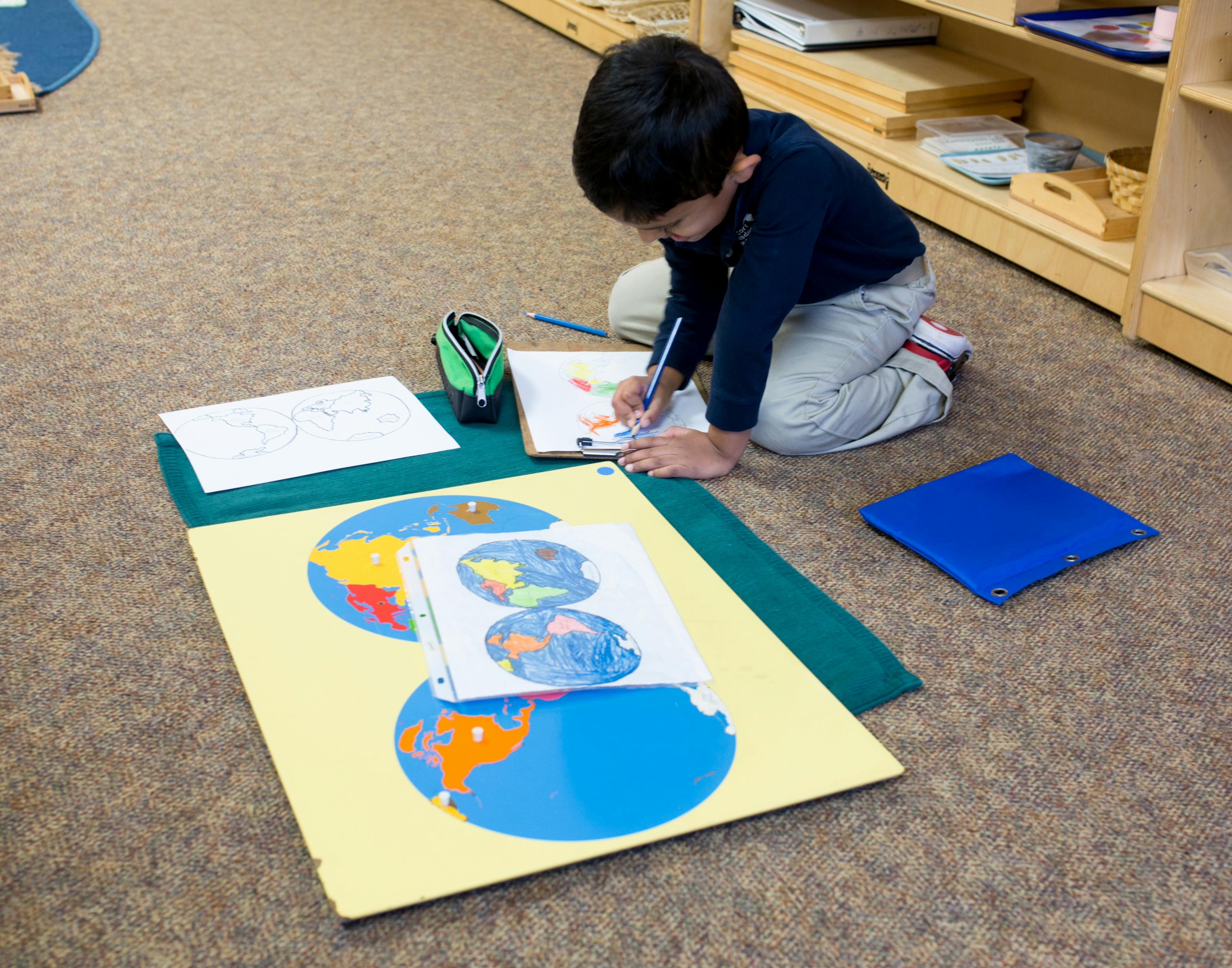 Child kneeling on floor drawing on paper with world map puzzle pieces nearby