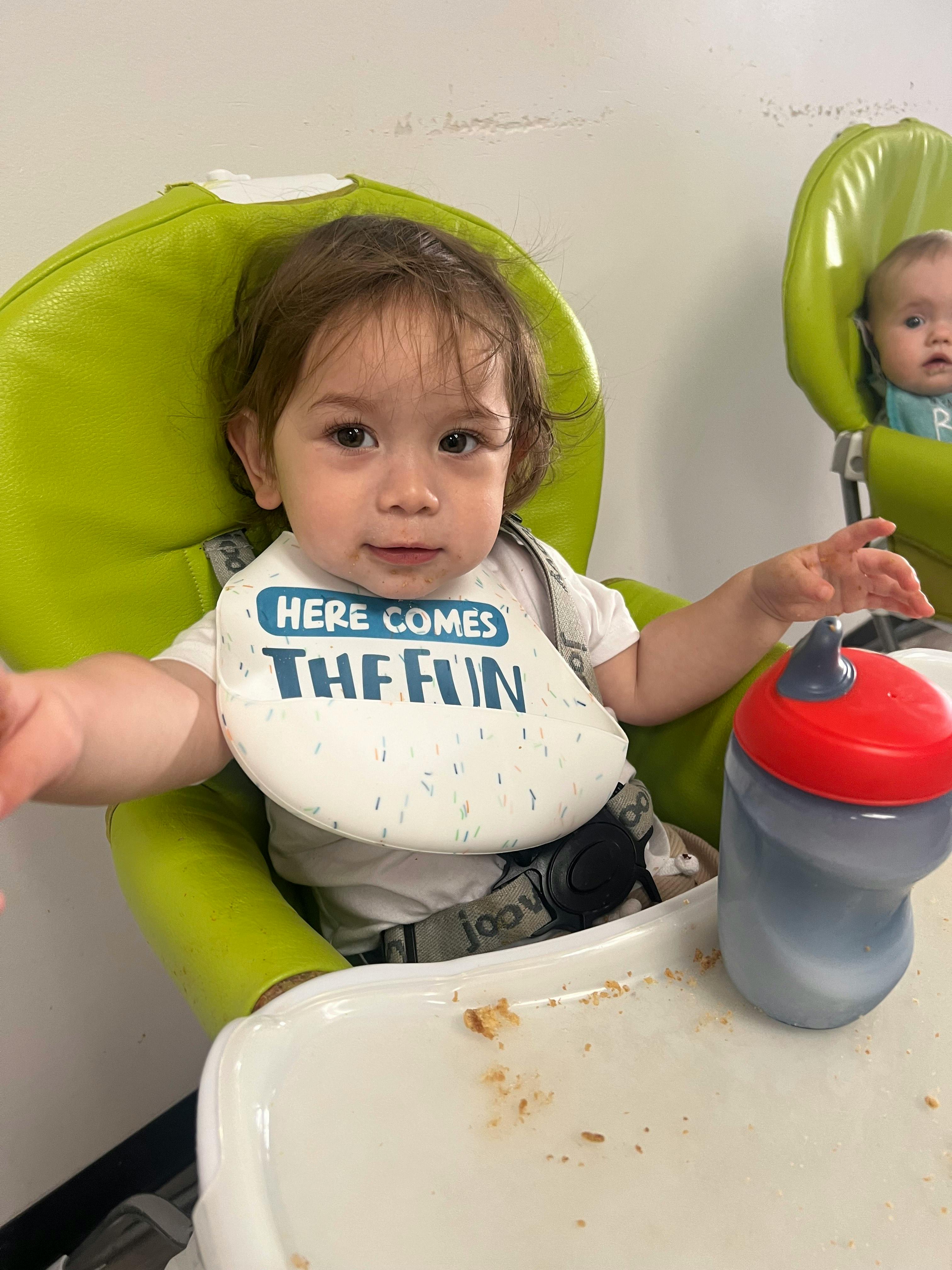 Child in a high chair wearing a bib that says "HERE COMES THE FUN"