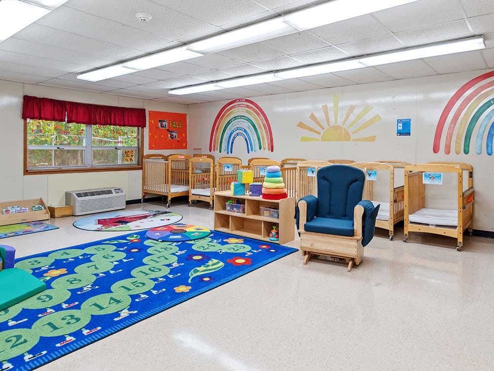 A colorful infant and toddler classroom with multiple cribs, rugs, and a rocking chair