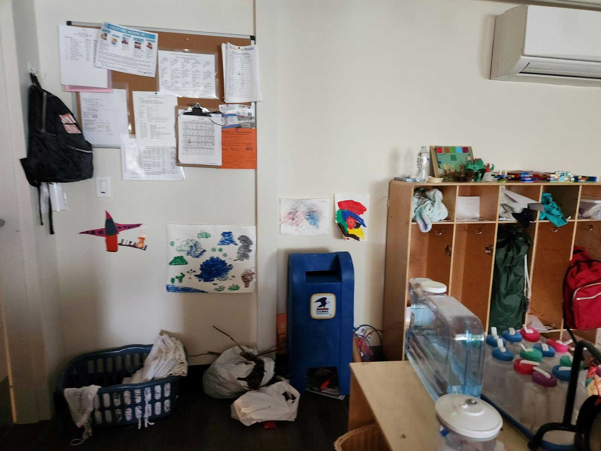 Messy classroom with a bulletin board, cubbies, and a blue recycling bin