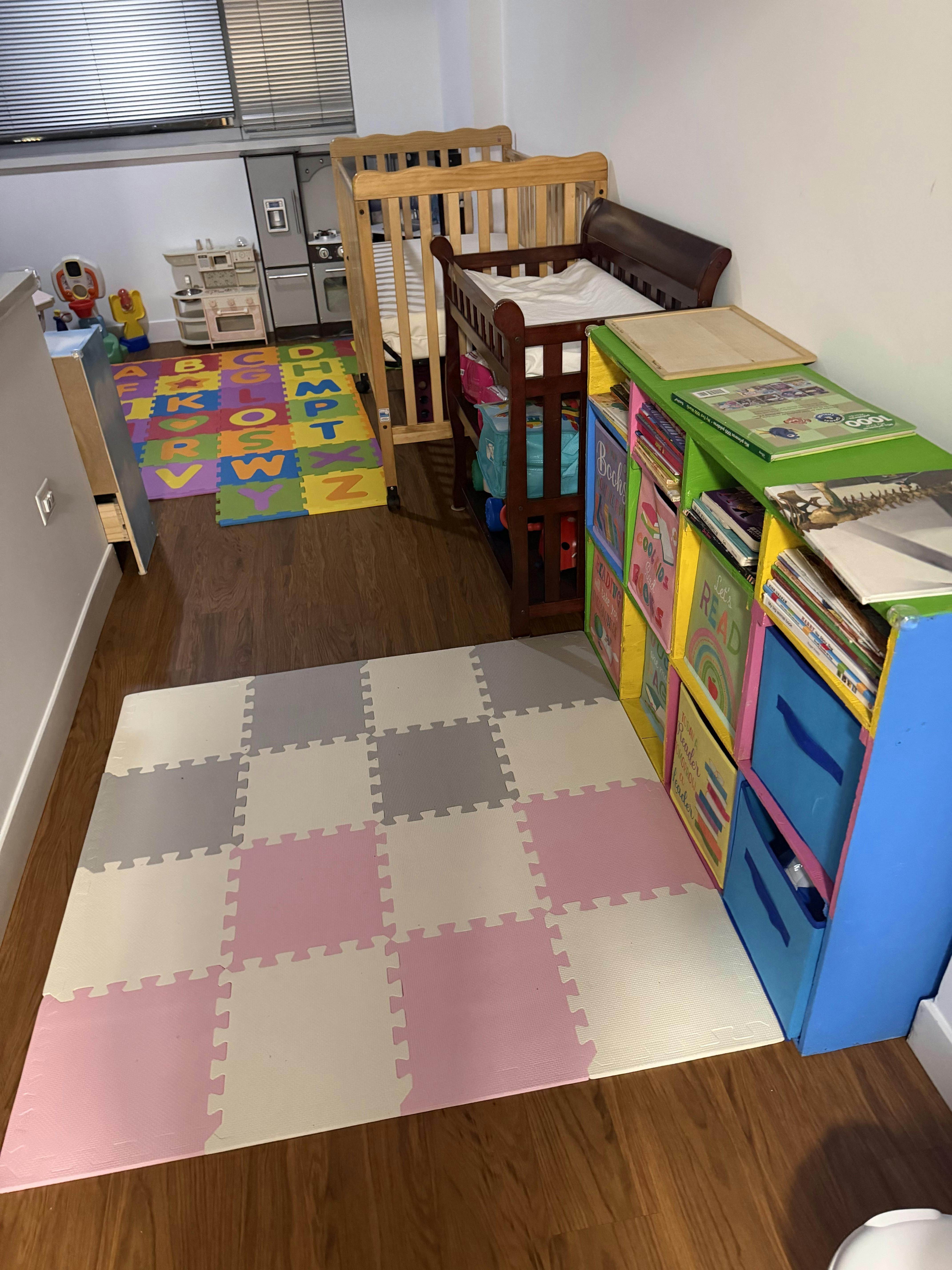 Child's play area with a crib, toy kitchen, storage shelves with books, and alphabet floor mats