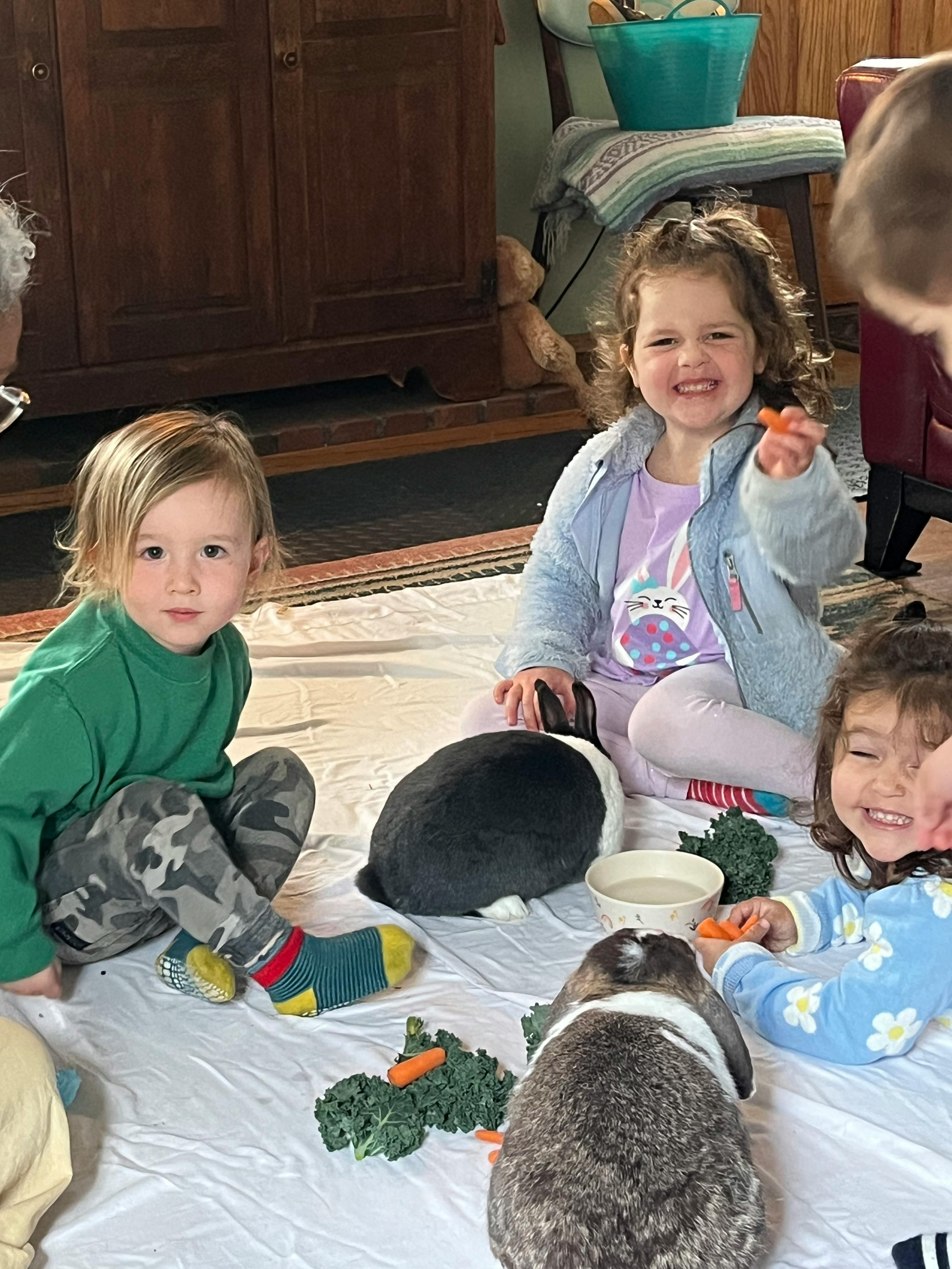 Three children sitting on the floor with two rabbits and vegetables