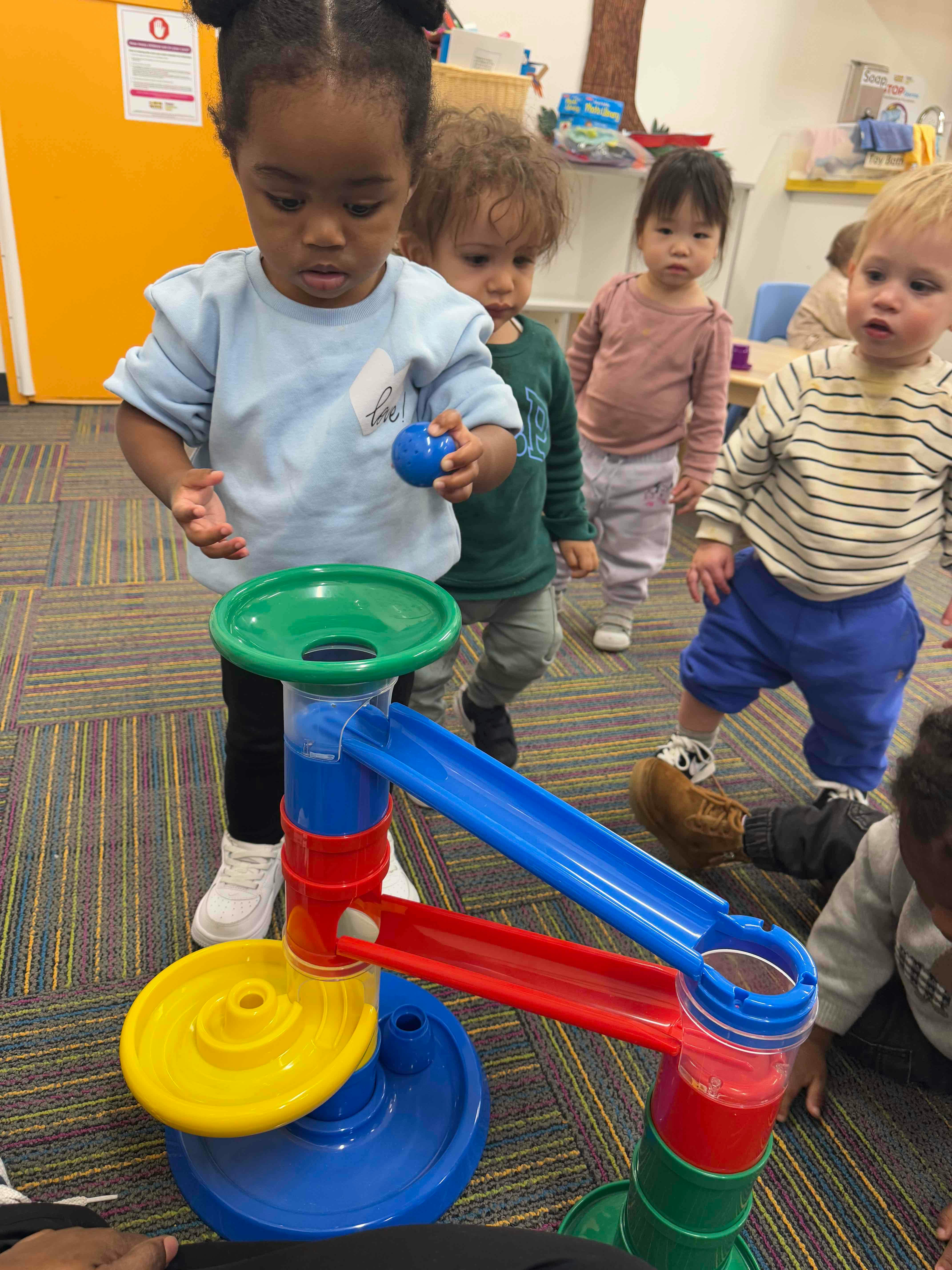 Child holding a ball over a colorful ball run toy with other children nearby