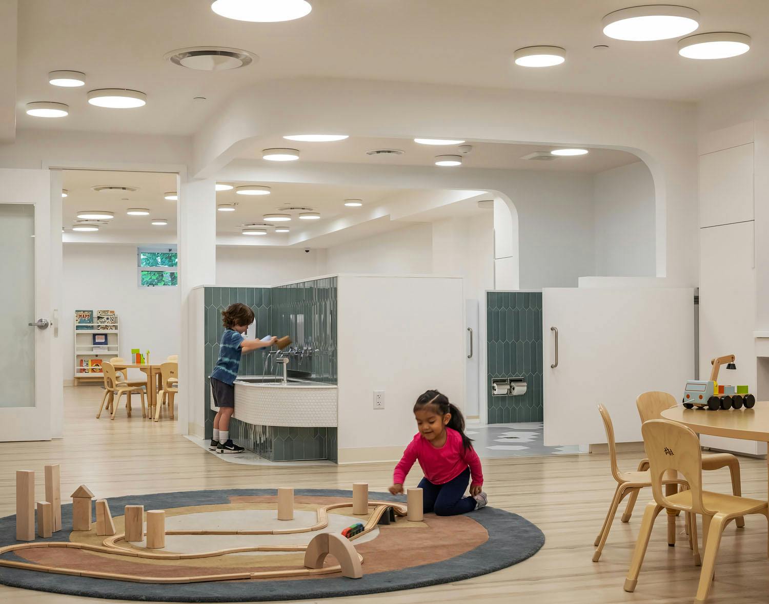 Child playing with blocks on a rug, another child at a sink in a classroom