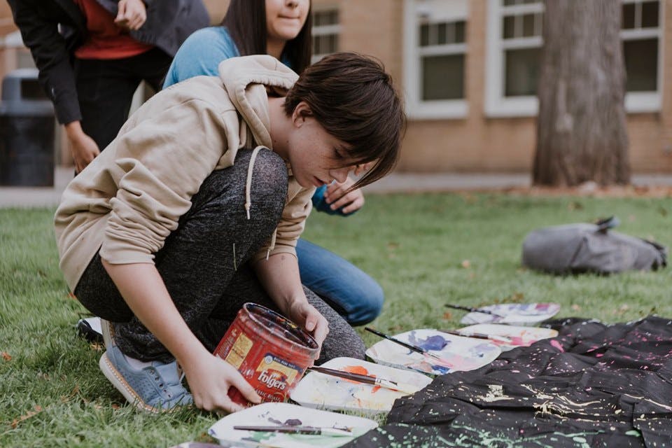 Child crouching on grass, holding a paint can over paint palettes
