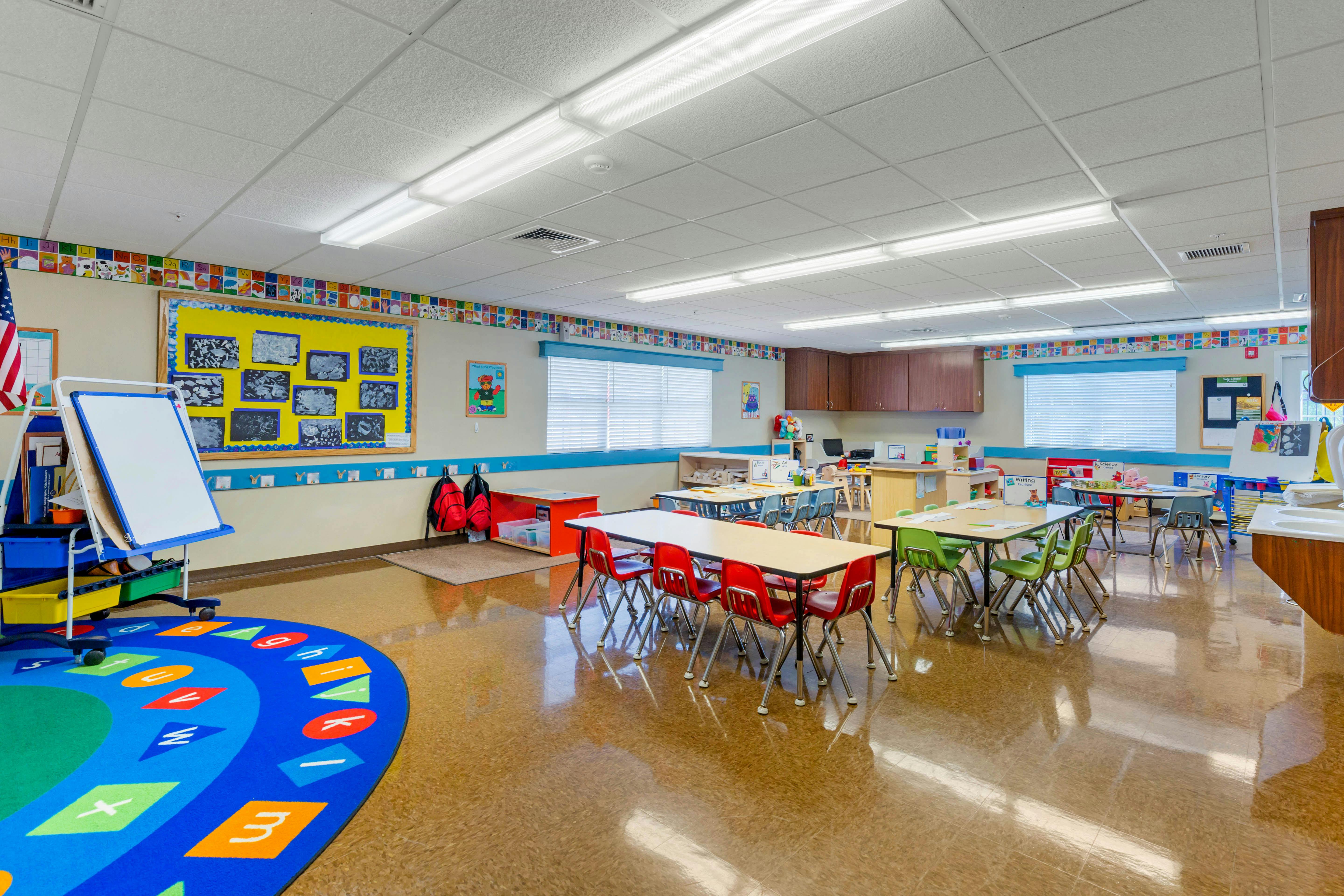Empty classroom with small tables and chairs, a whiteboard, and a colorful rug