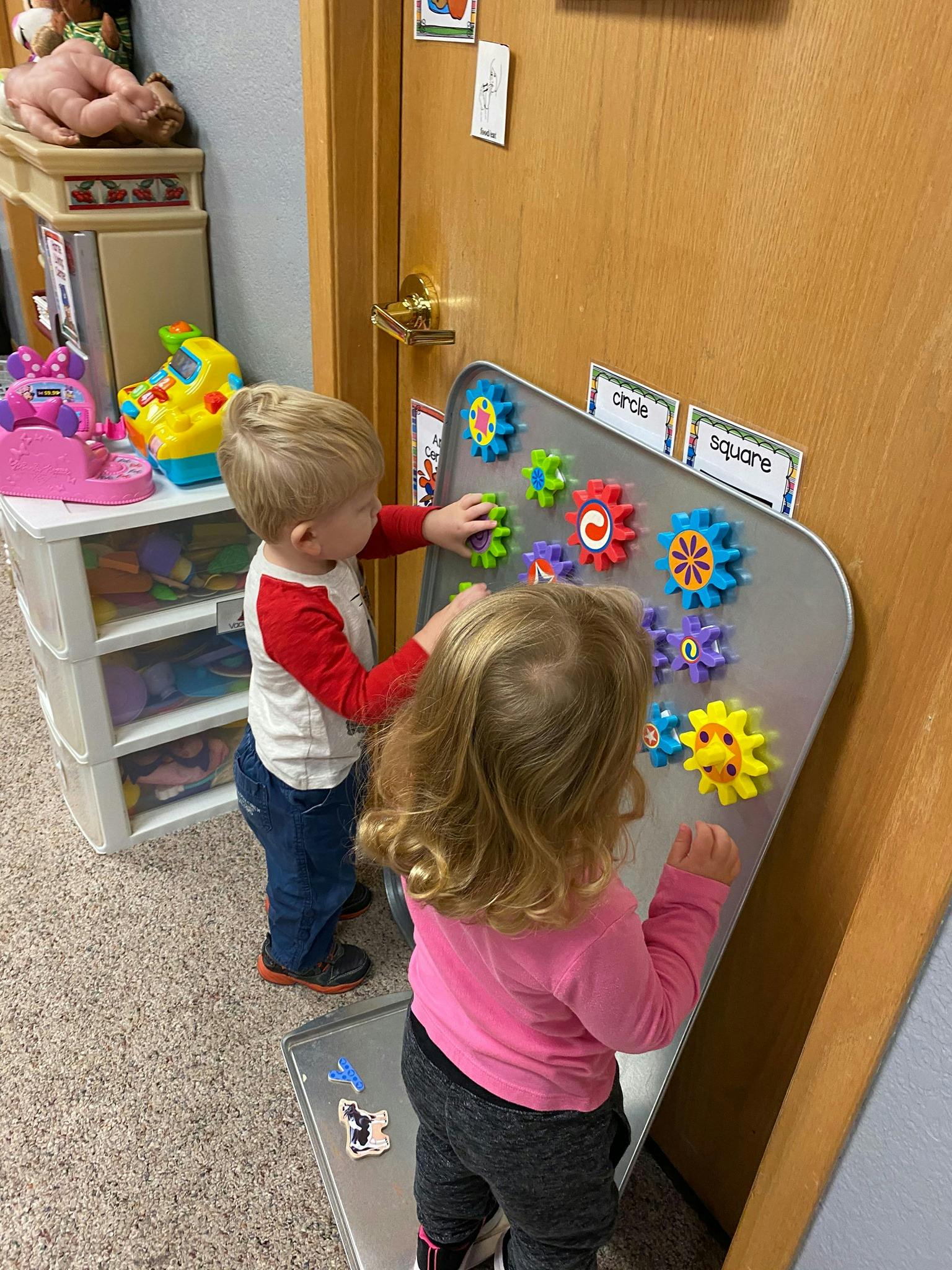 Two children playing with magnetic gears on a metal board in a classroom