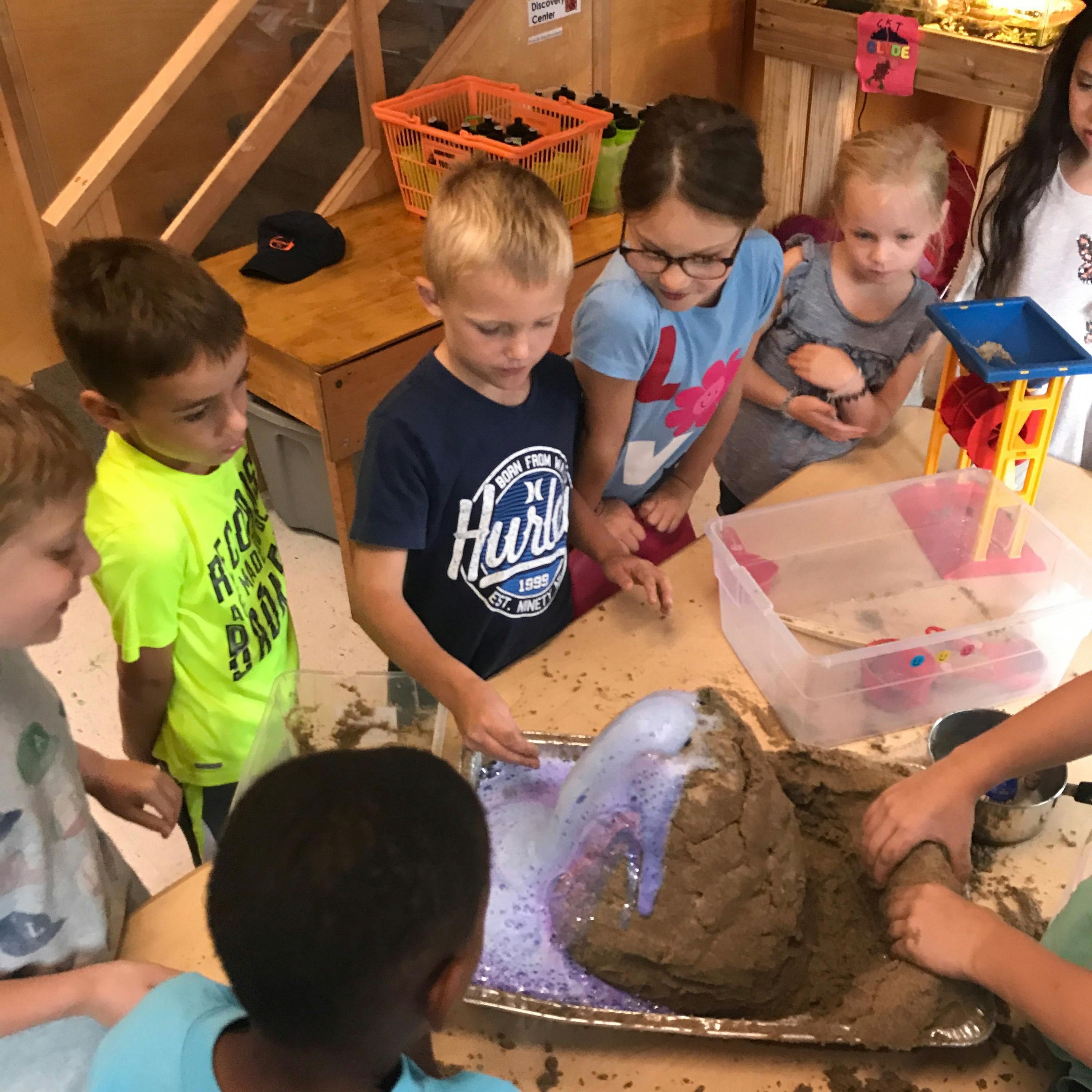 Children gathered around a table, watching a science experiment with a volcano