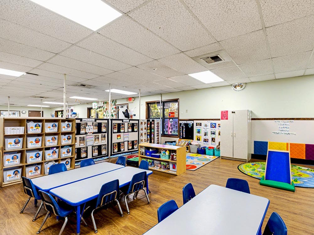 Empty classroom with tables, chairs, storage shelves, and play rugs