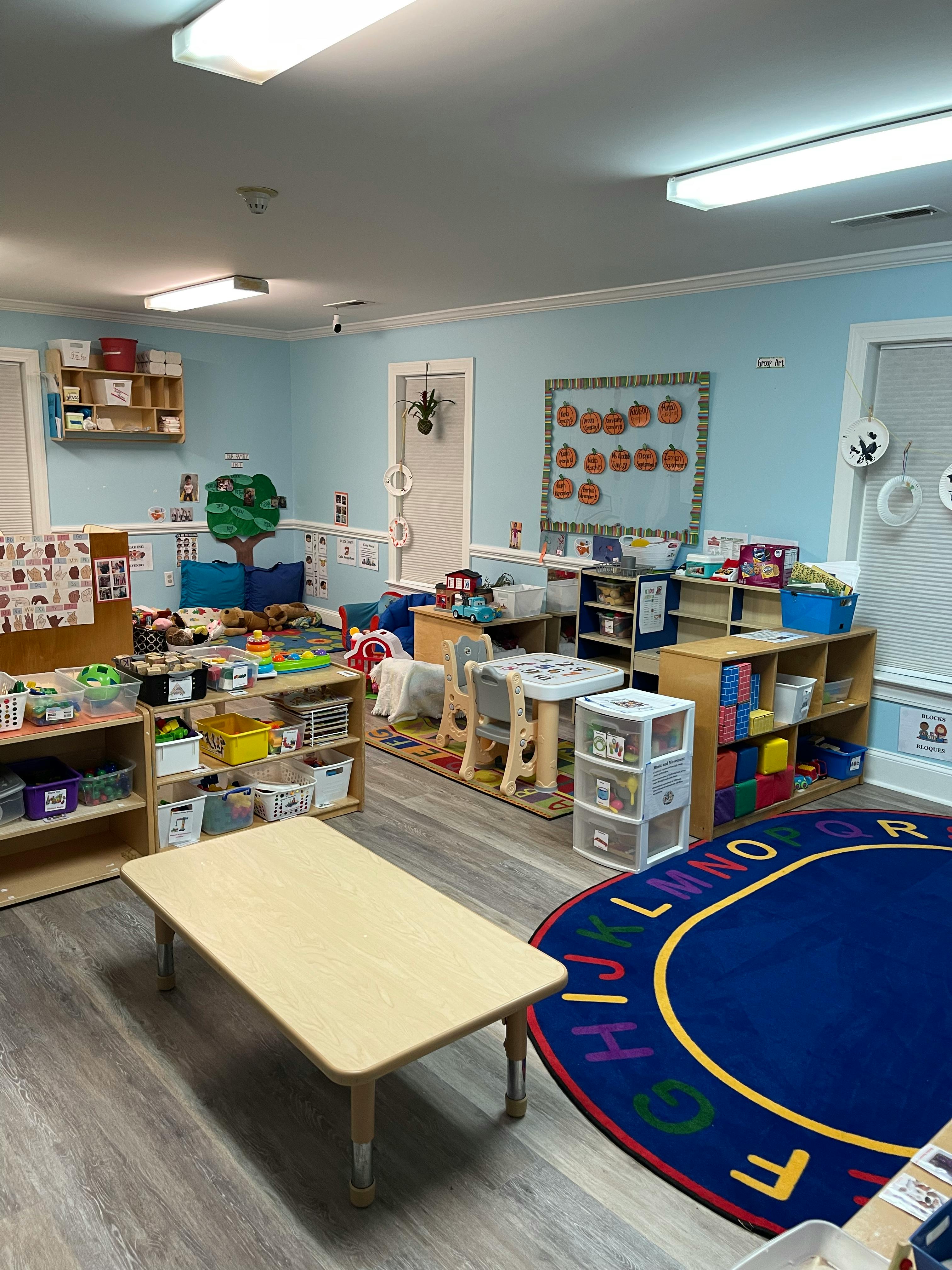 Classroom with small tables, chairs, shelves, and a blue alphabet rug