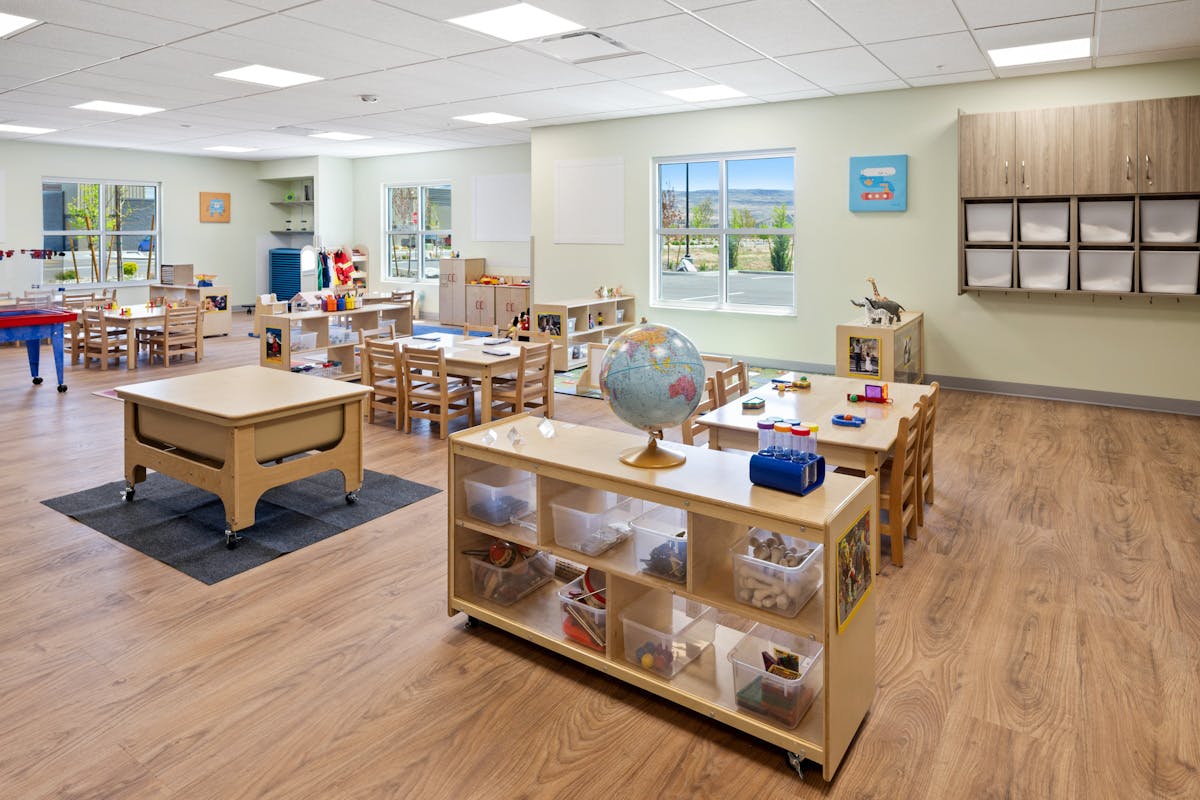 An empty classroom with small tables and chairs, shelves with toys, and a globe