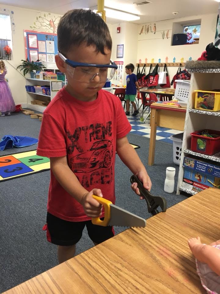 Child in safety glasses holding toy saw and wrench in a classroom
