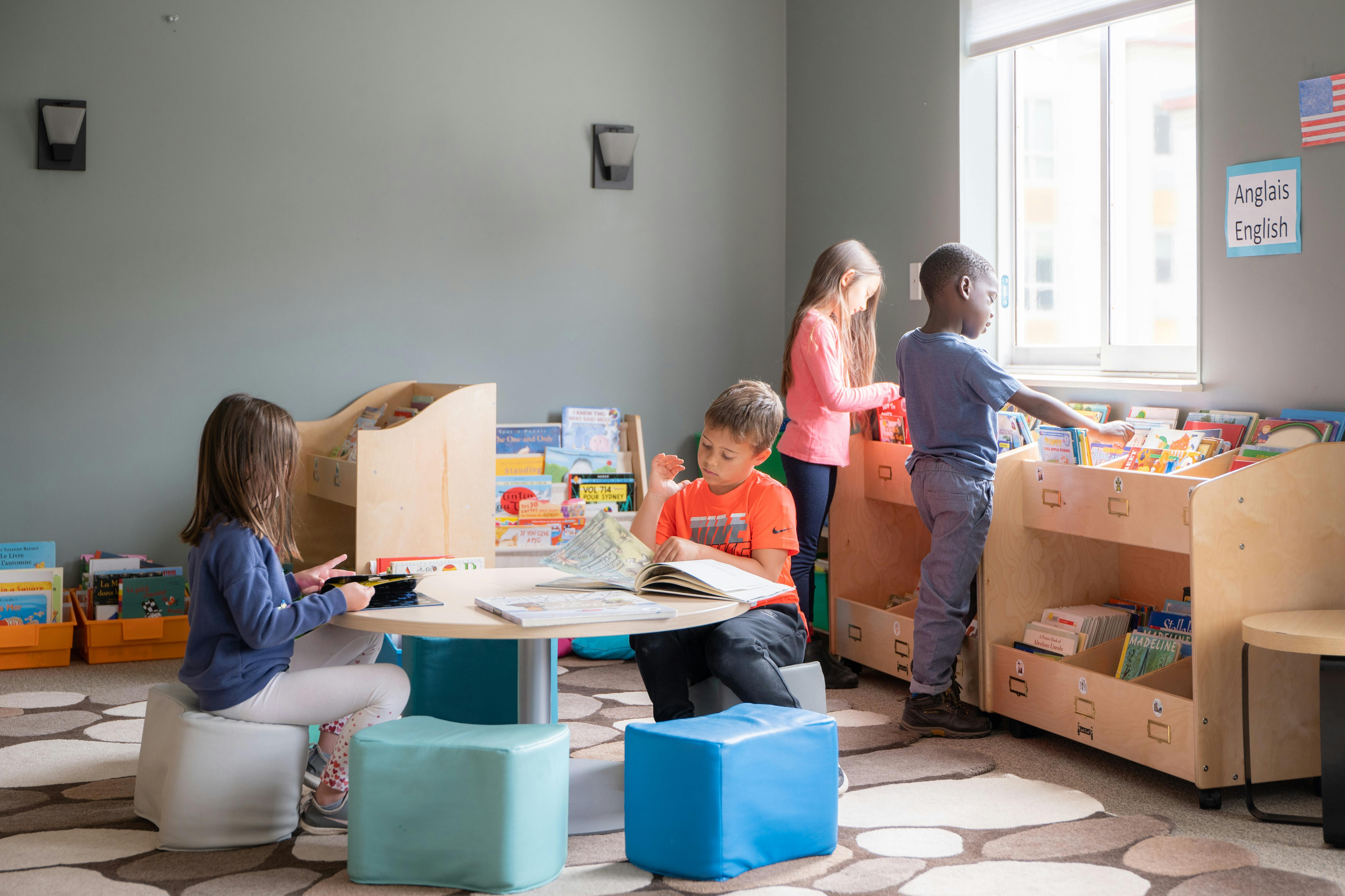 Children in a classroom, some reading at a table, others looking at shelves