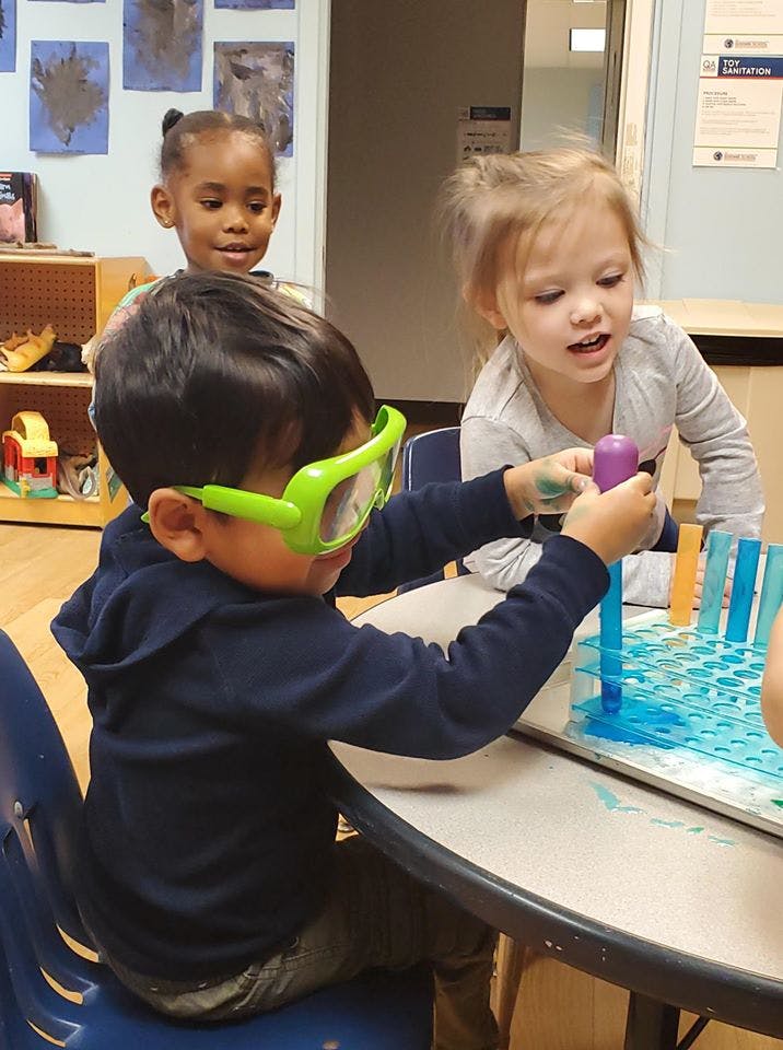 Child in green goggles playing with tubes and liquid at a table with other children