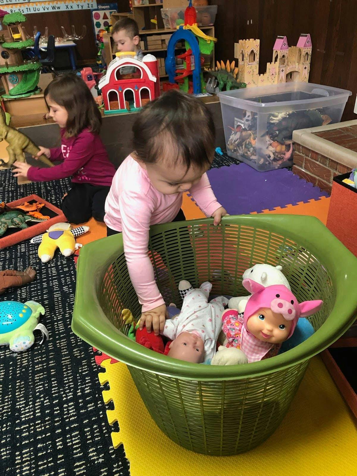 Child reaching into a plastic basket containing toy dolls in a playroom