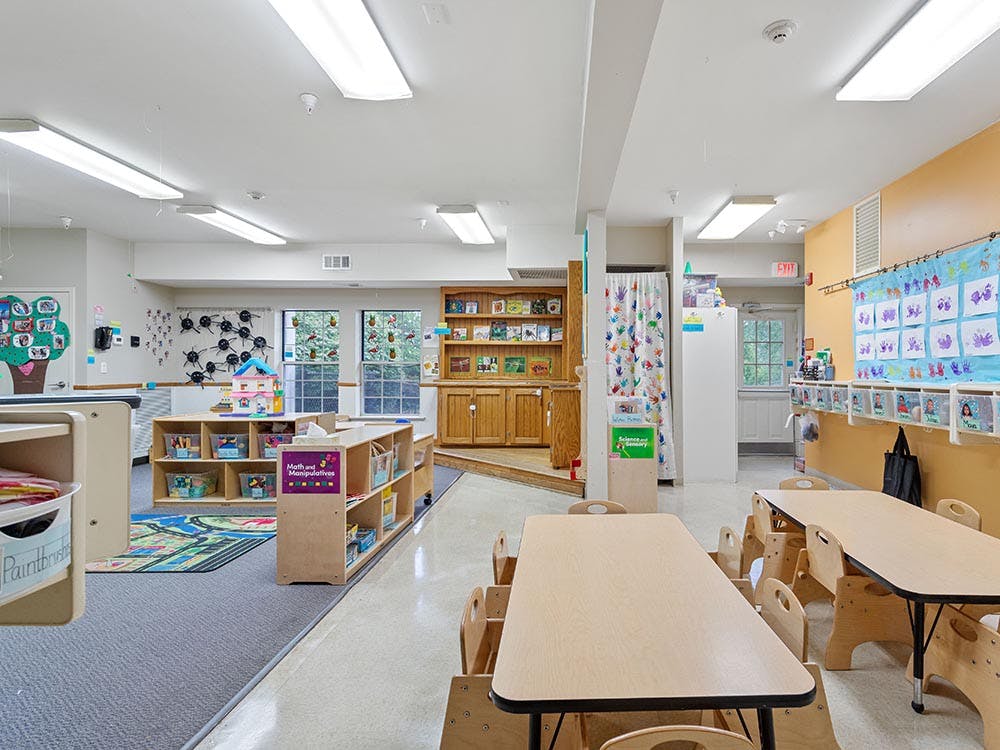 Empty classroom with small tables and chairs, shelves with toys, and decorated walls