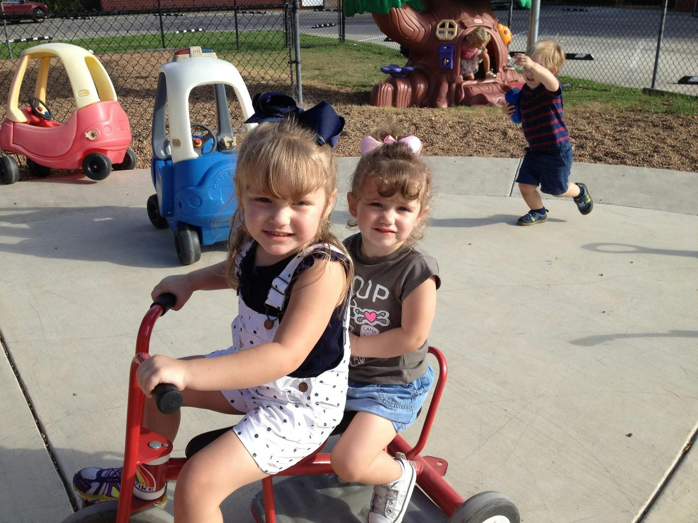 Two children on a tricycle with another child playing in the background