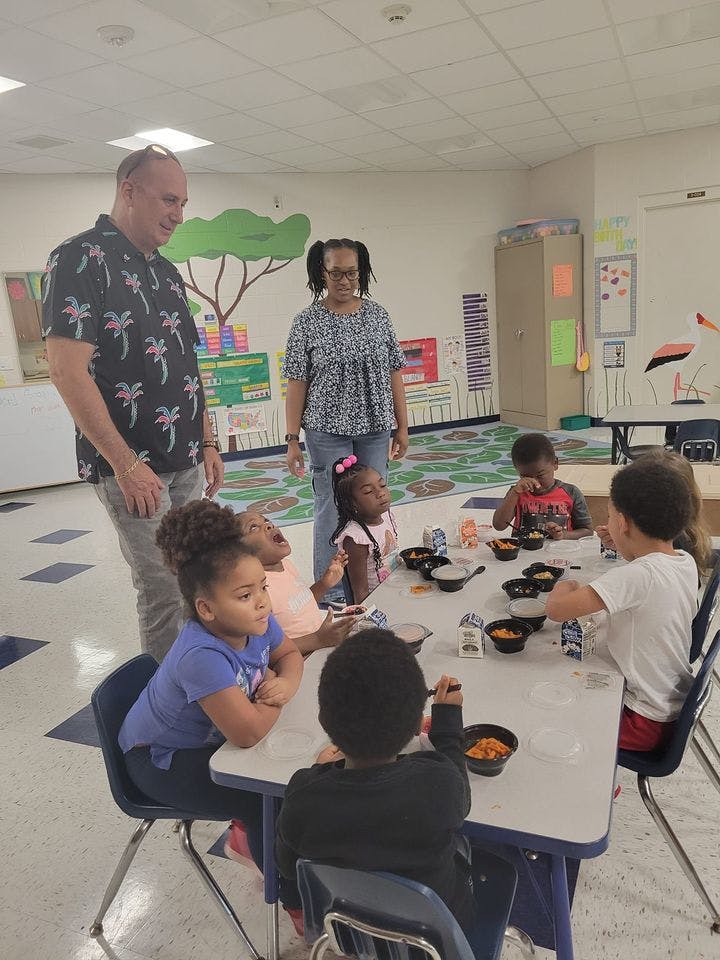 Two adults standing near children eating at a table in a classroom