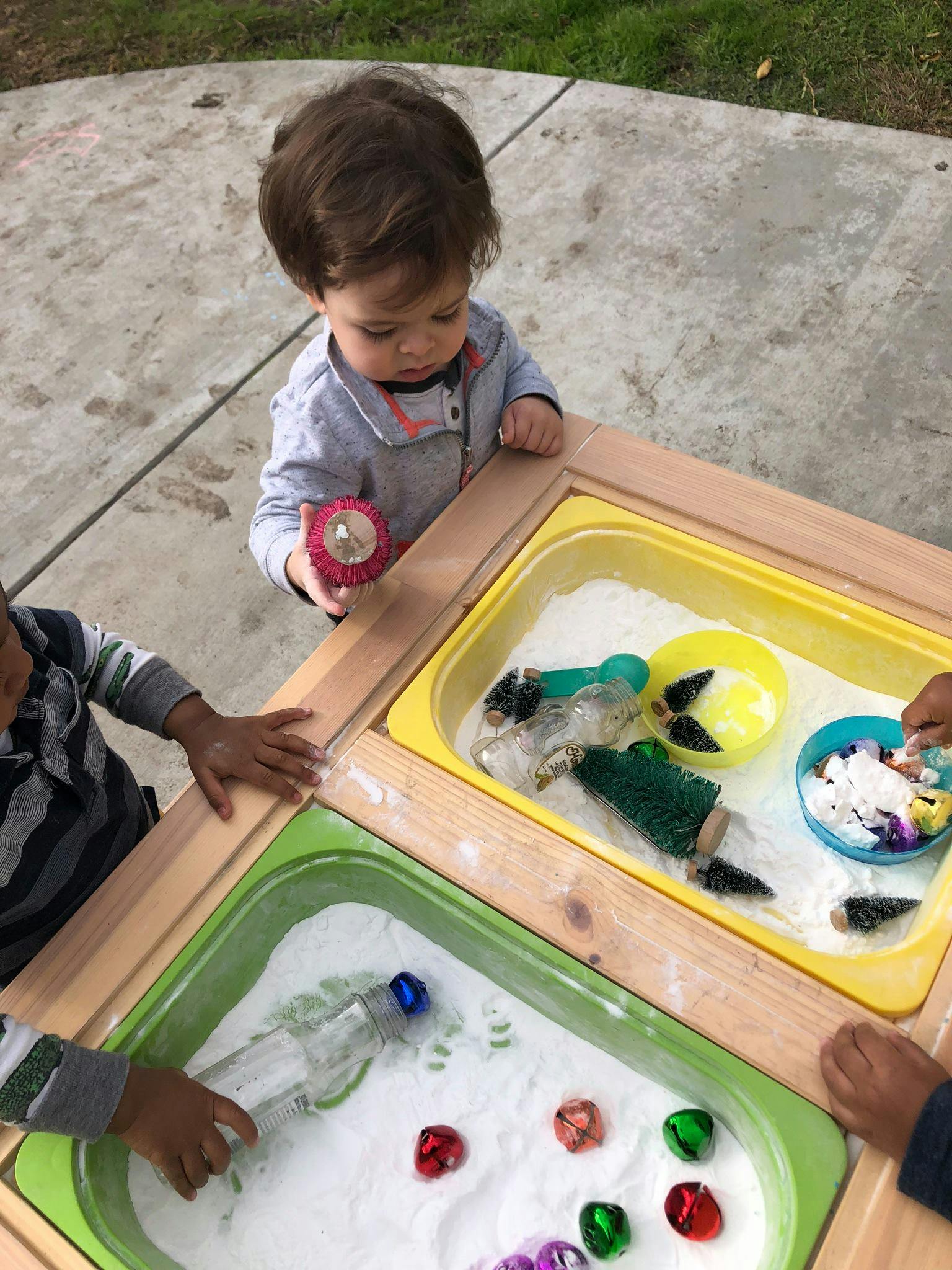 Child holding a red toy over a sensory table with white sand and toys