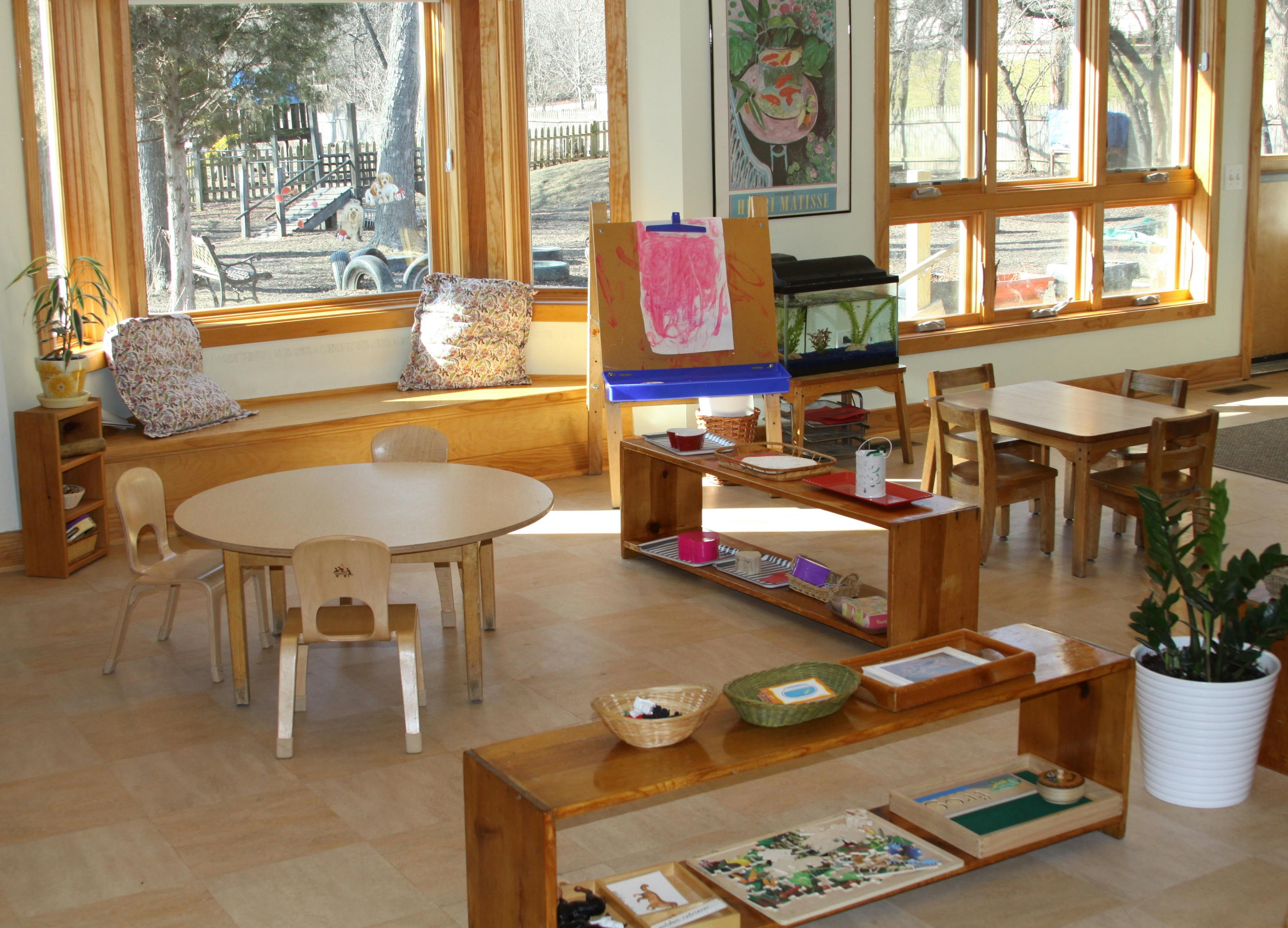 Classroom with small wooden tables and chairs, shelves, and large windows
