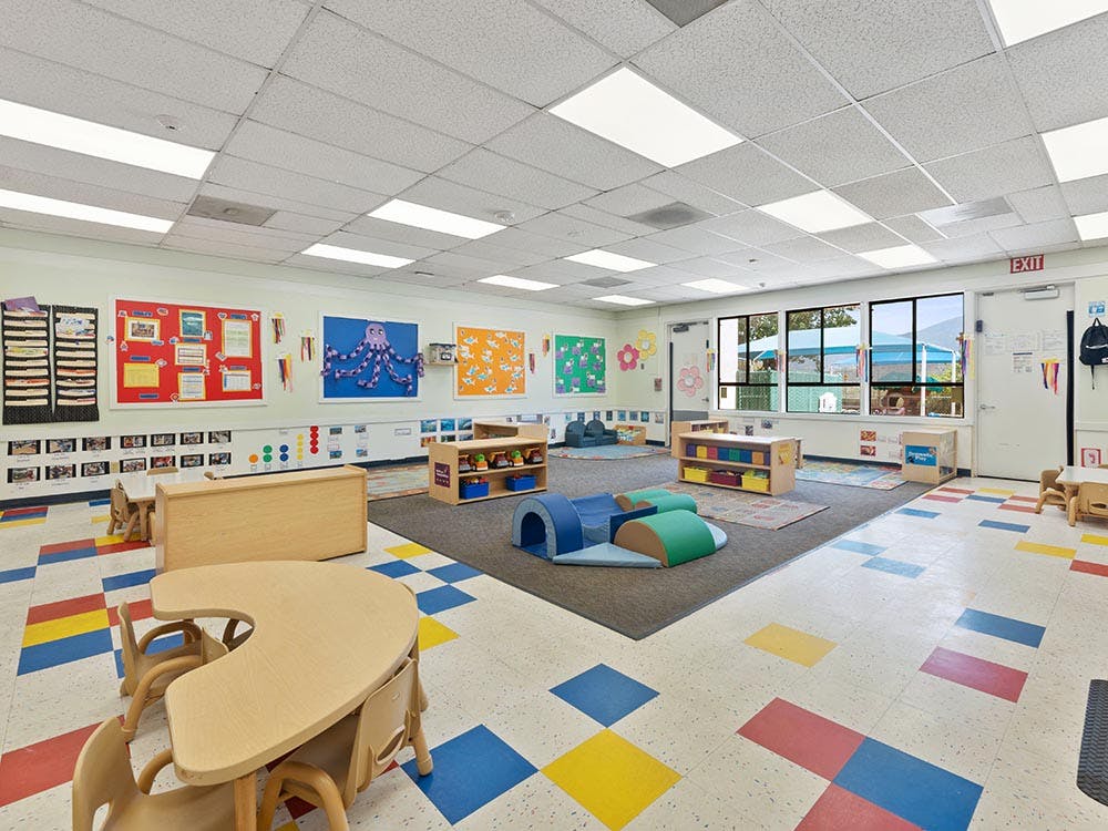 Colorful classroom with small tables and chairs, play mats, and wall decorations