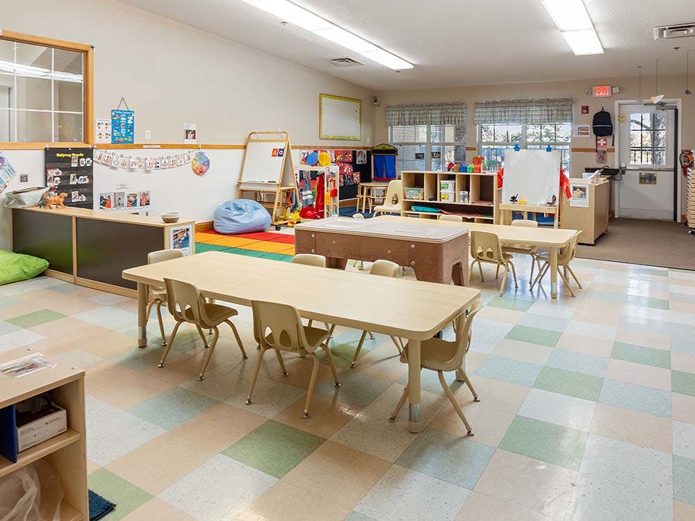 Classroom with tables and chairs for children, whiteboards, and colorful play rug