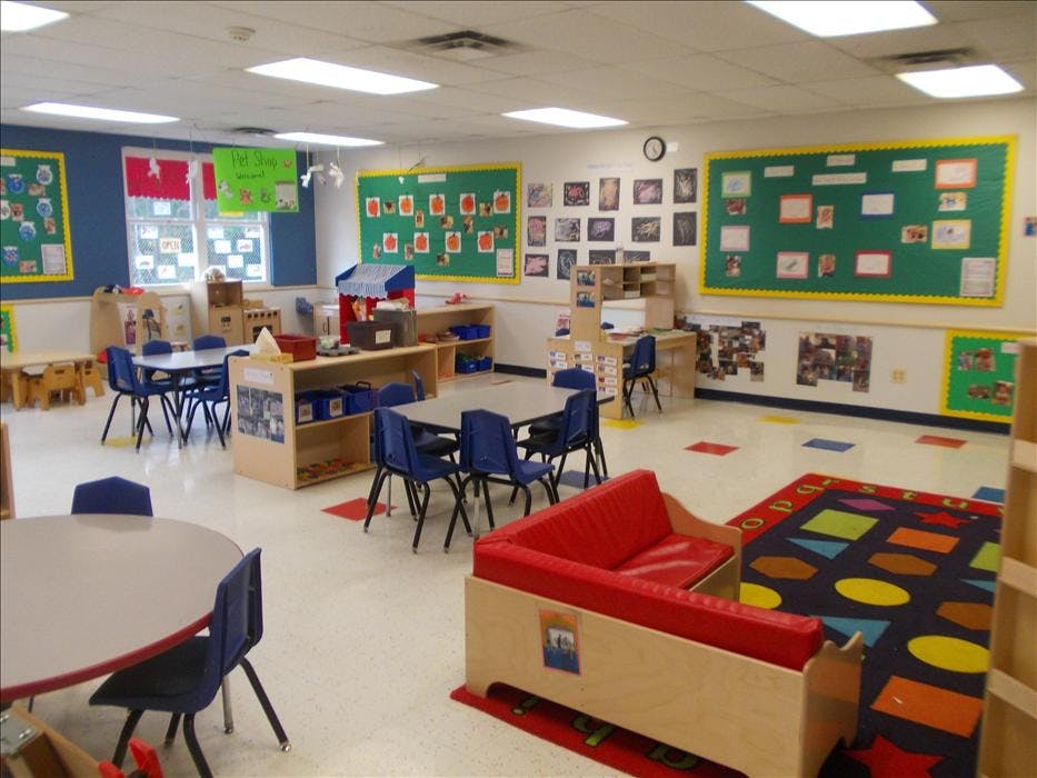 Empty classroom with small tables and chairs, play kitchen, and bulletin boards