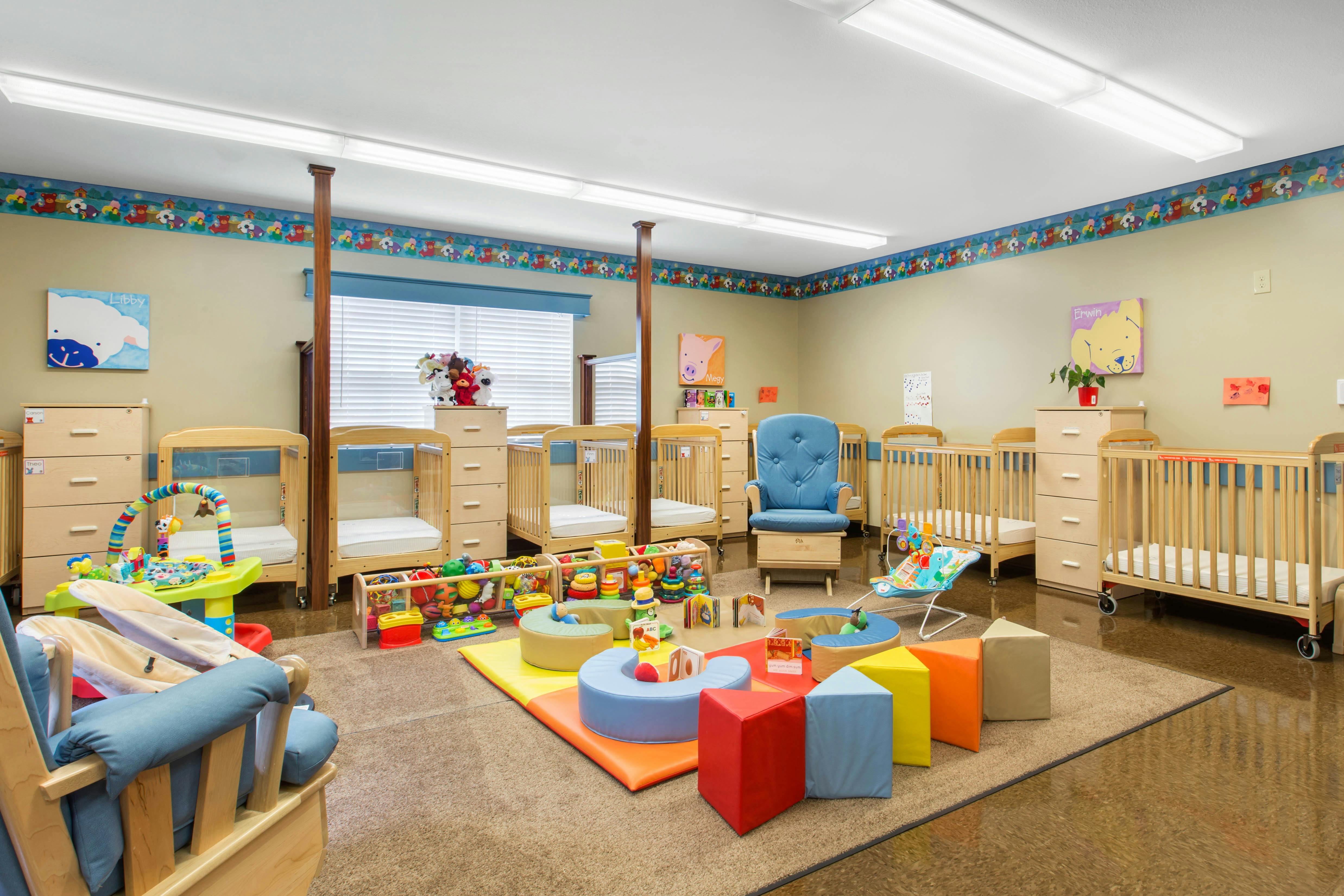 A brightly lit nursery room with multiple cribs, a play mat with colorful toys, and a blue rocking chair