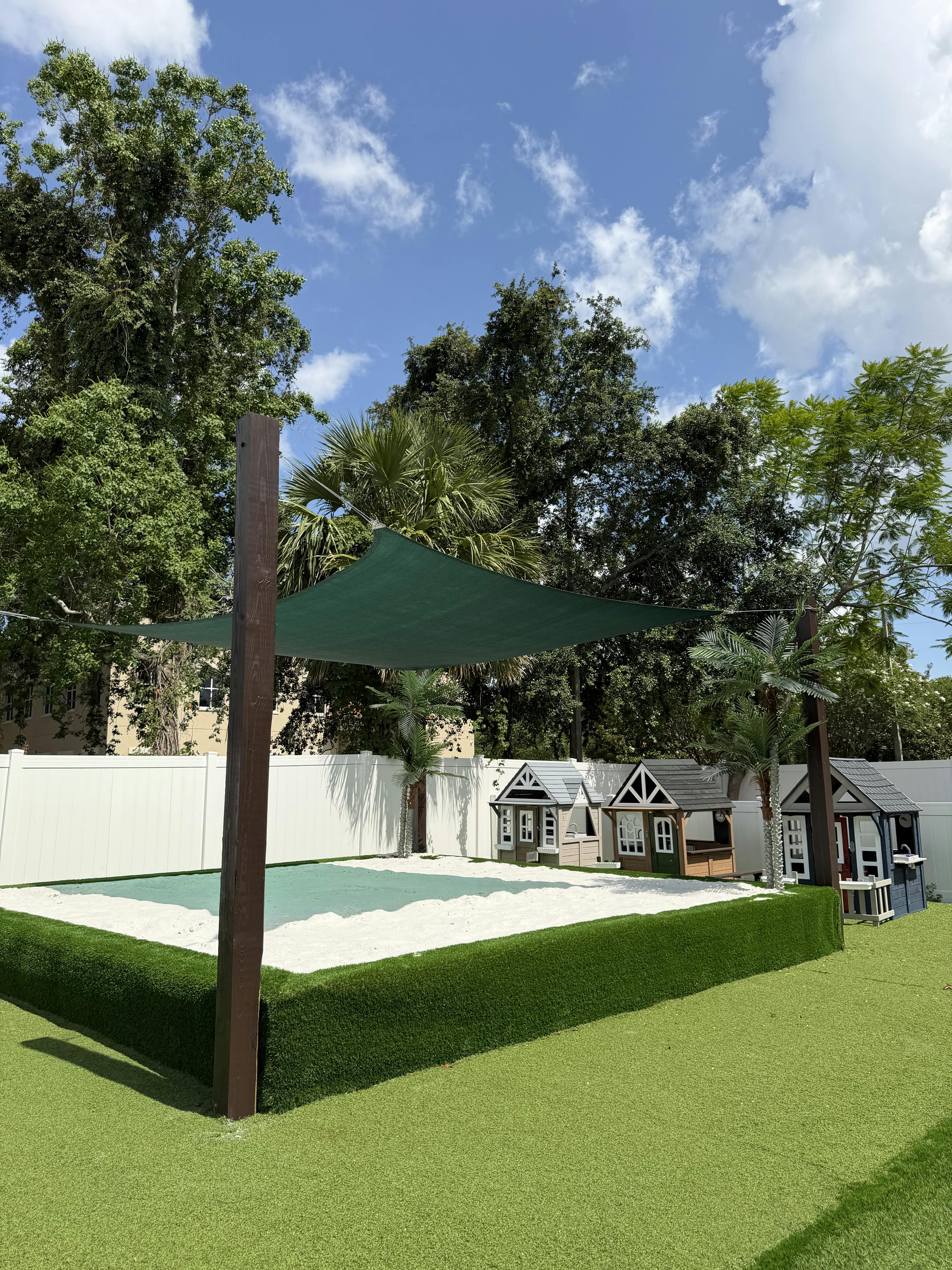 Sandbox with a green shade sail and small playhouses in a backyard