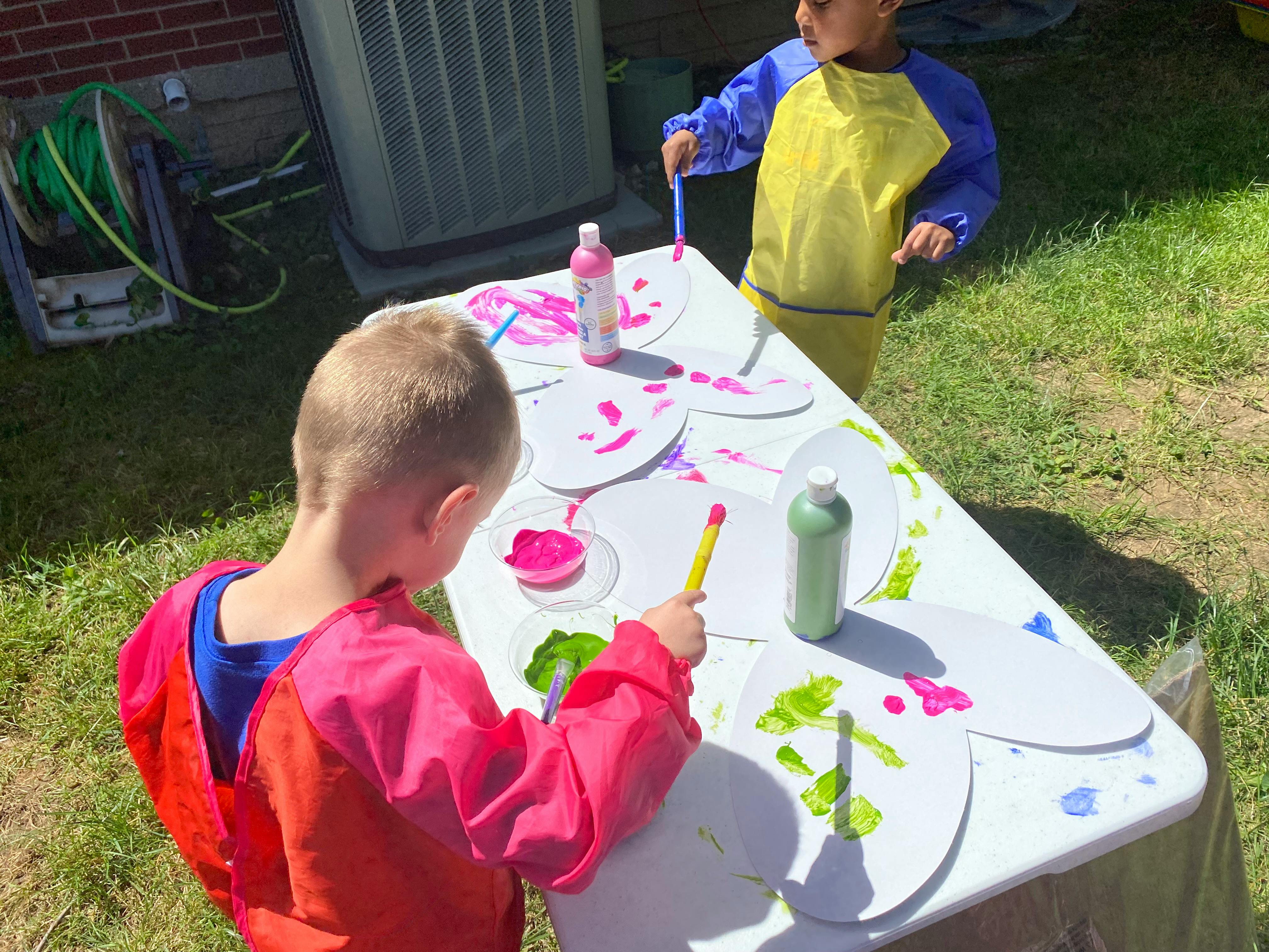 Two children wearing art smocks painting paper butterfly shapes at an outdoor table