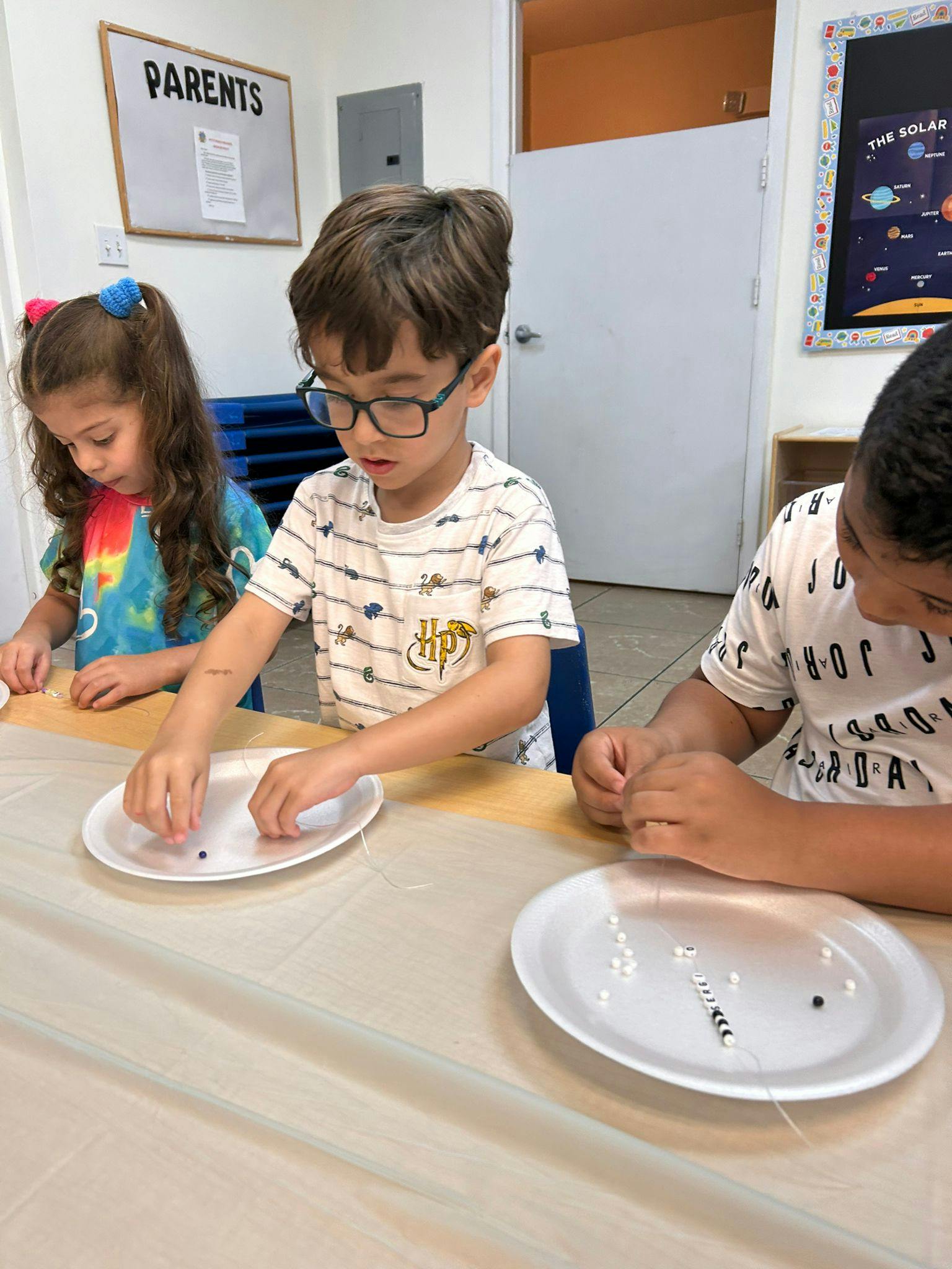 Three children making beaded jewelry at a table