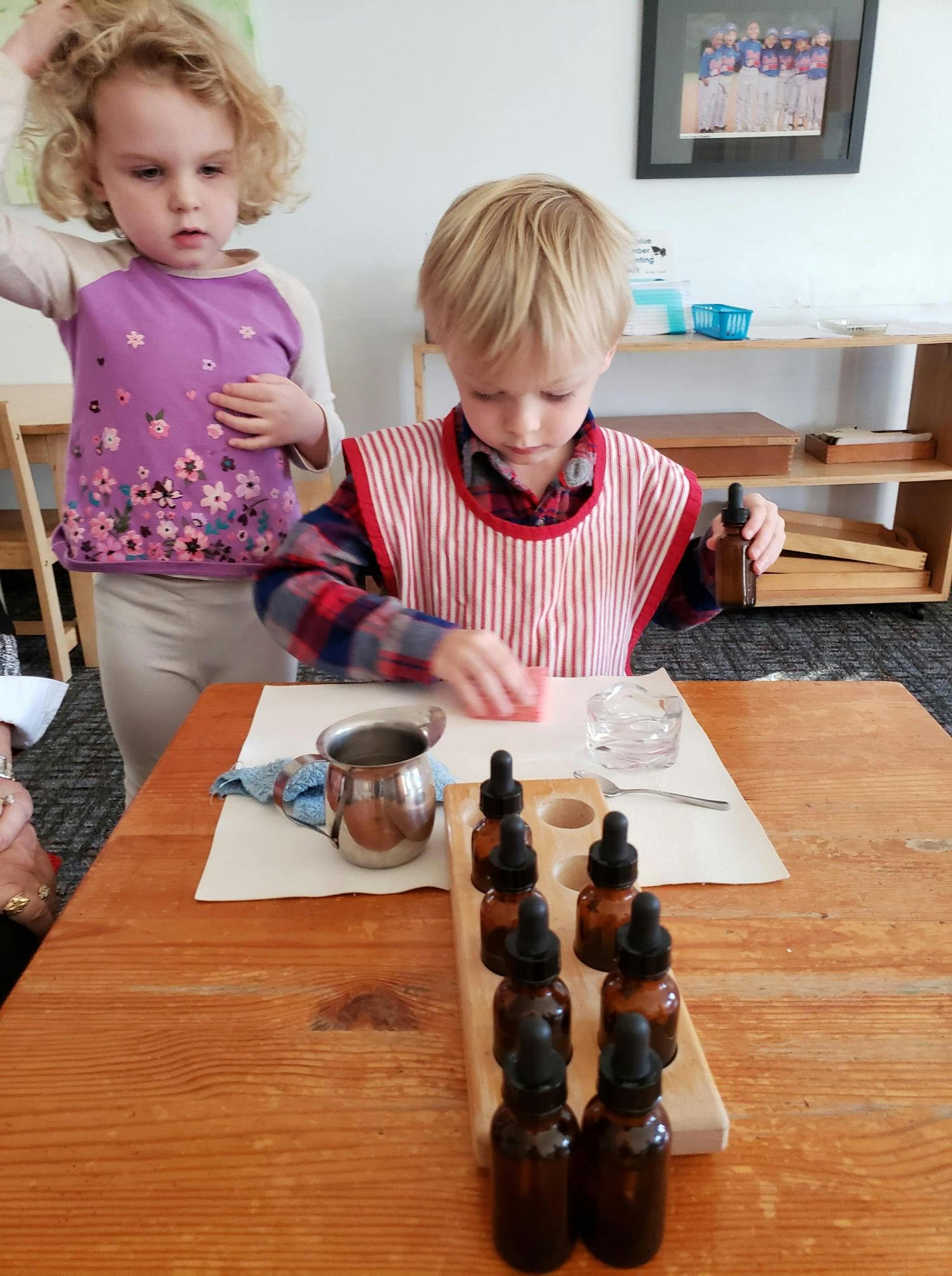Child in a striped apron working with dropper bottles at a table, another child nearby