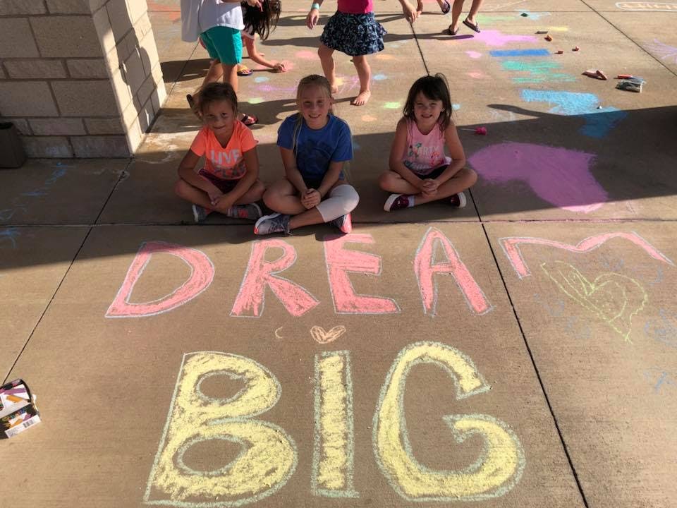 Three children sitting on pavement with "DREAM BIG" written in chalk