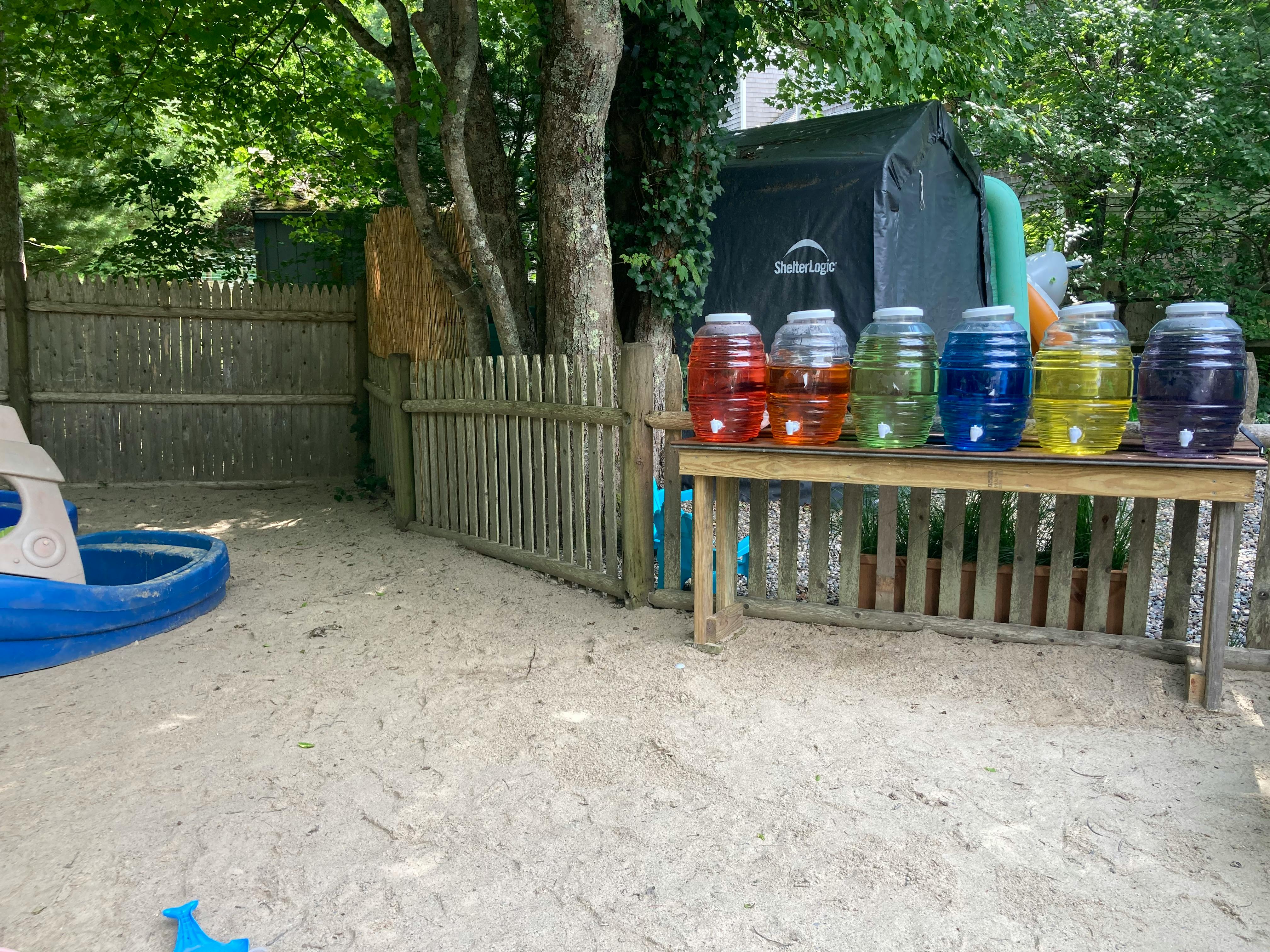 Six large jars of colorful liquid on a wooden table outdoors