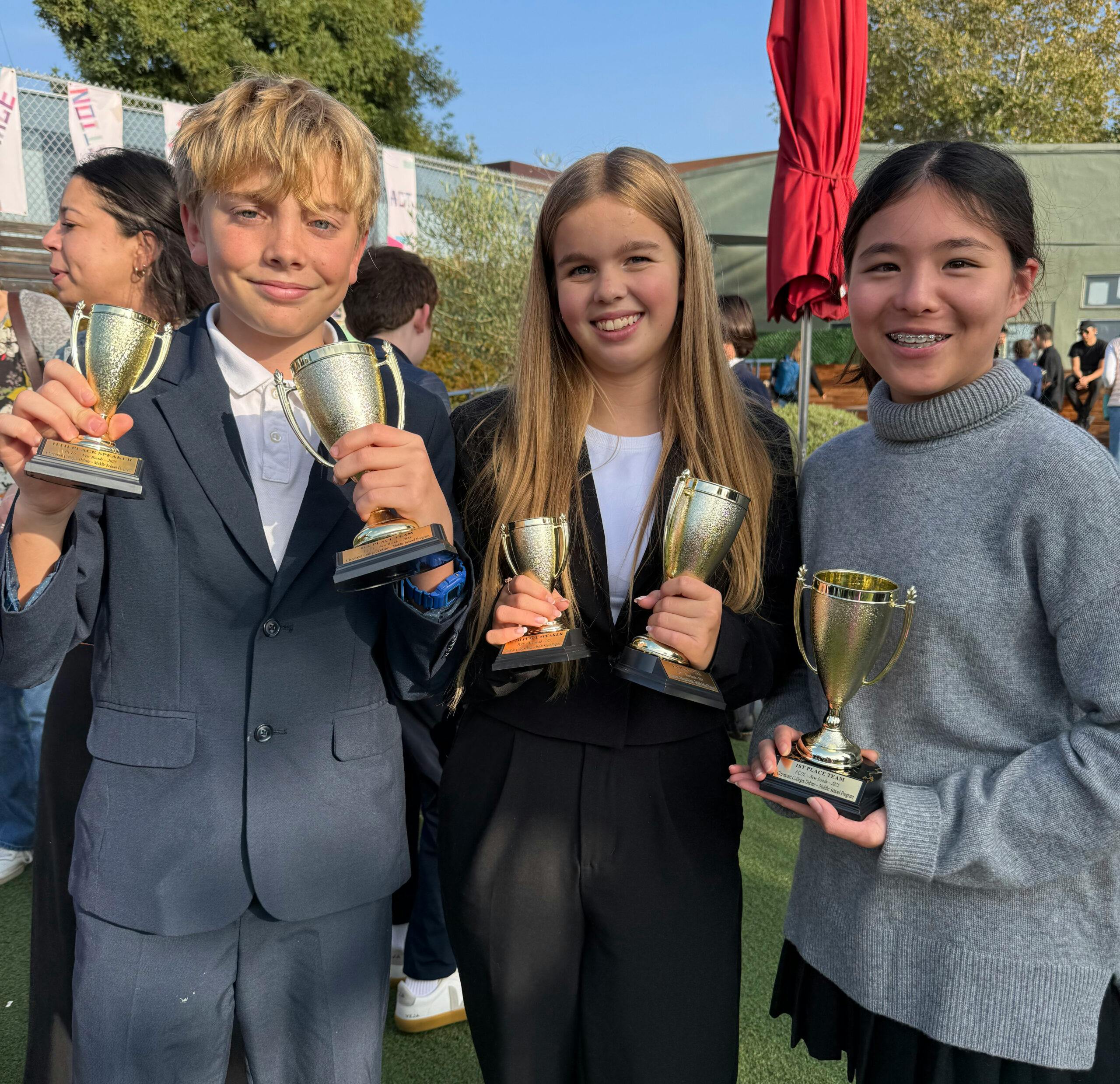 Three teens holding up debate trophies, two wearing blazers, one in a gray sweater