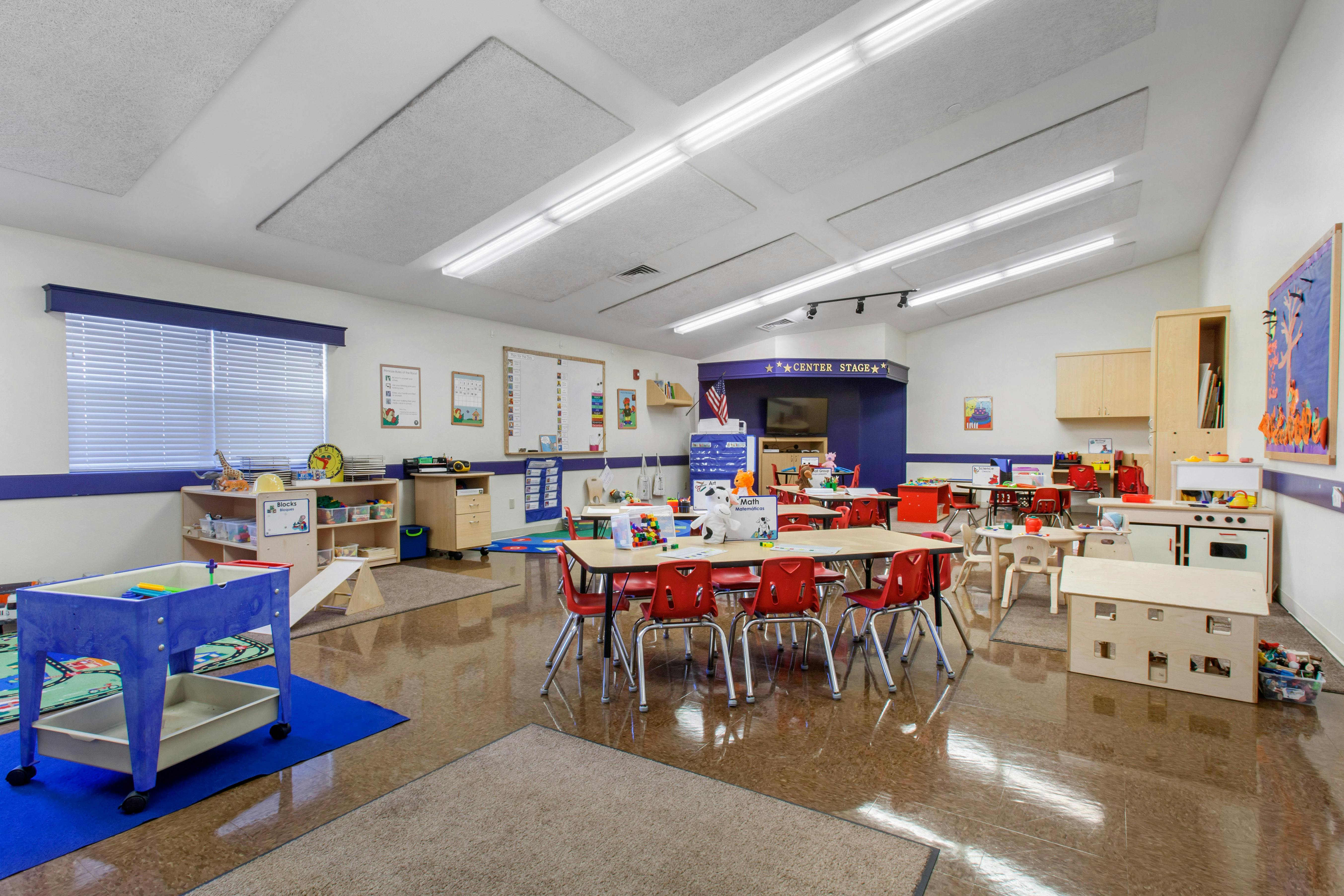 A colorful classroom with small red chairs and tables, a play kitchen, and toys